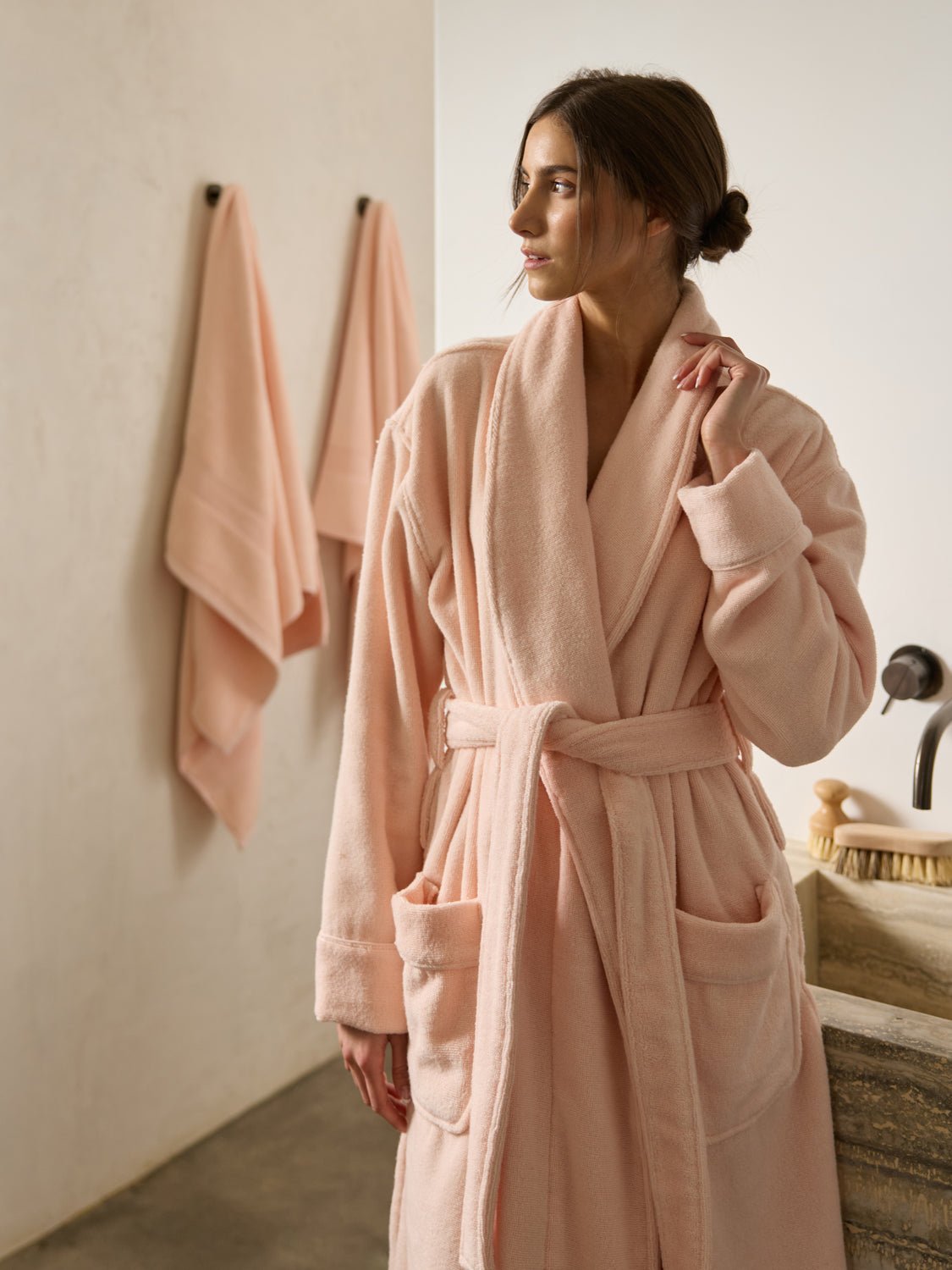 A woman stands in a bathroom wearing a Cozy Earth Luxe Bath Robe in light pink, featuring large pockets and tied at the waist. Two matching towels hang on the wall behind her. 