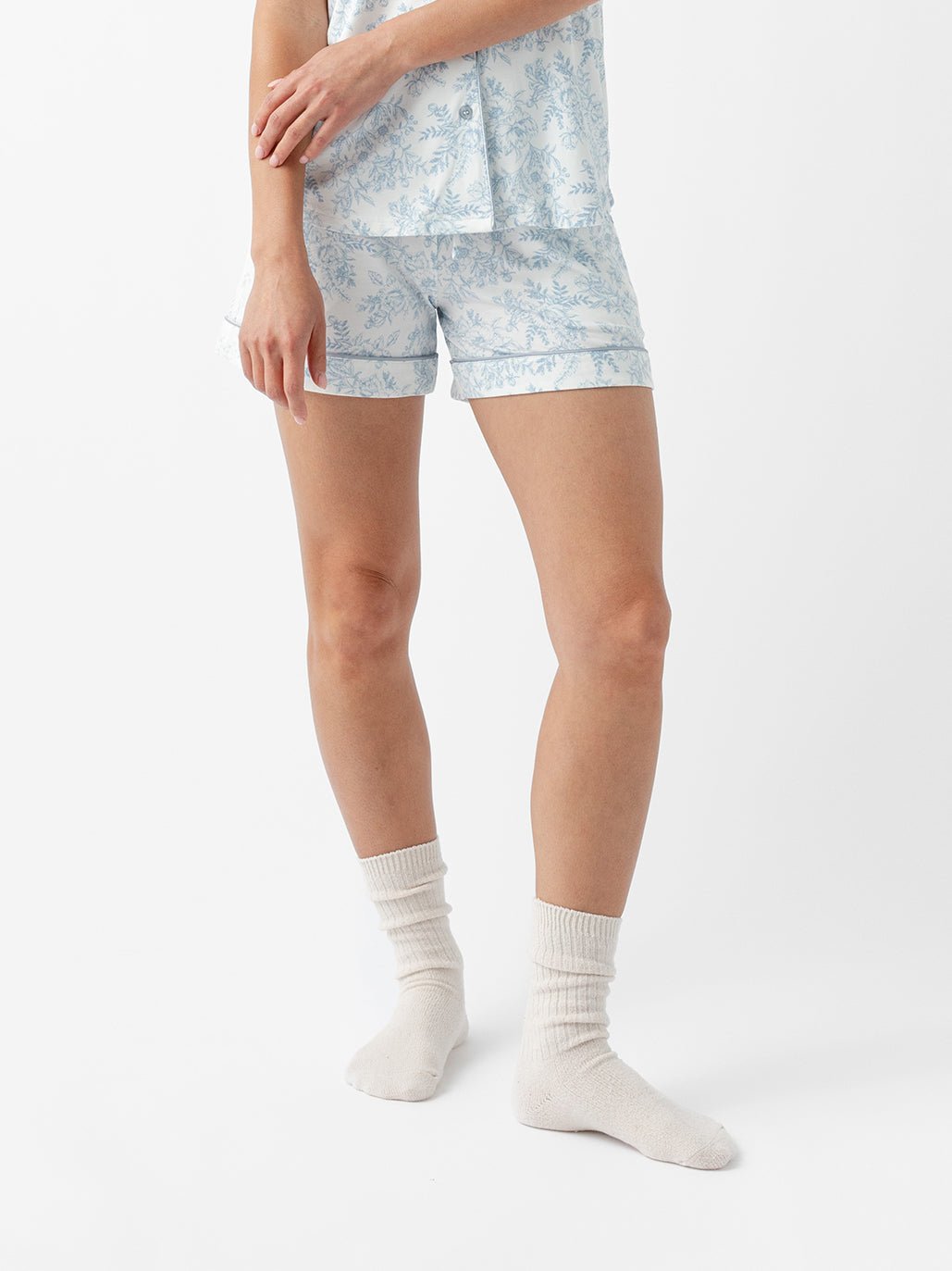 A person in Cozy Earth Women's Bamboo Stretch-Knit Pajama Short, featuring a light blue floral design with shorts and shirt, stands against a white background. Paired with cream socks, their face is not visible. 