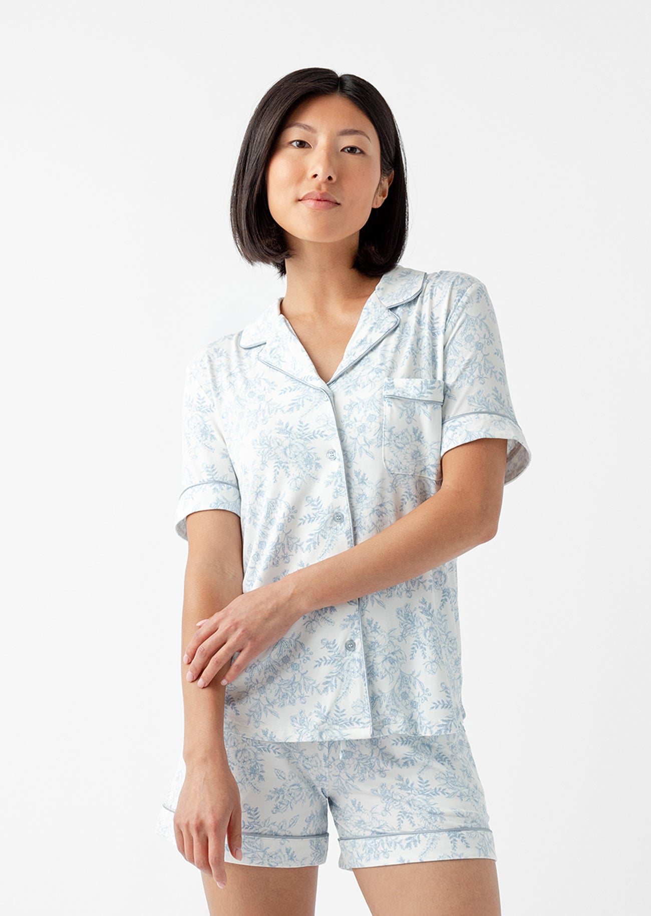 A person with shoulder-length black hair is wearing a Sky Toile pajama set, featuring the Women's Bamboo Stretch-Knit Short Sleeve Top by Cozy Earth, paired with shorts. They stand against a plain white background. |Color:Sky Toile