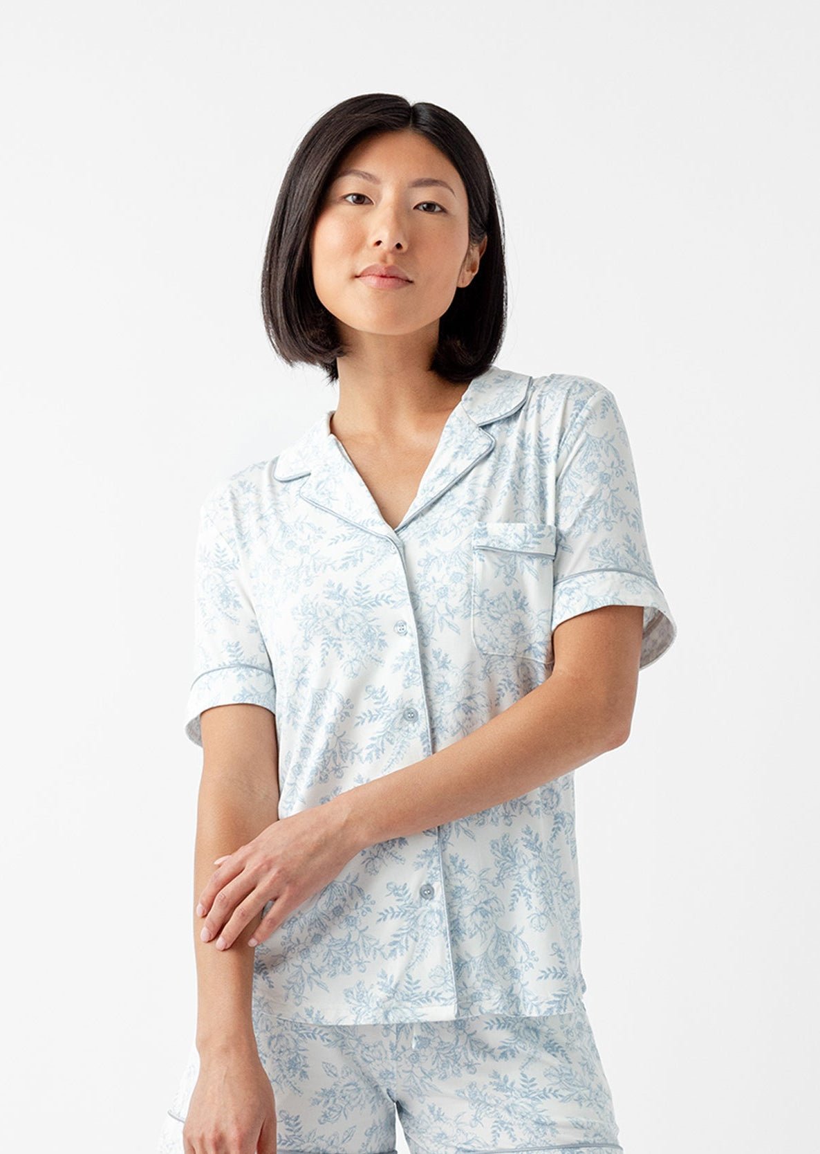 A person with shoulder-length black hair wears the Women's Bamboo Stretch-Knit Short Sleeve & Pant Pajama Set by Cozy Earth, featuring a light blue color and subtle floral pattern. They stand against a white background, looking slightly to the side. 
