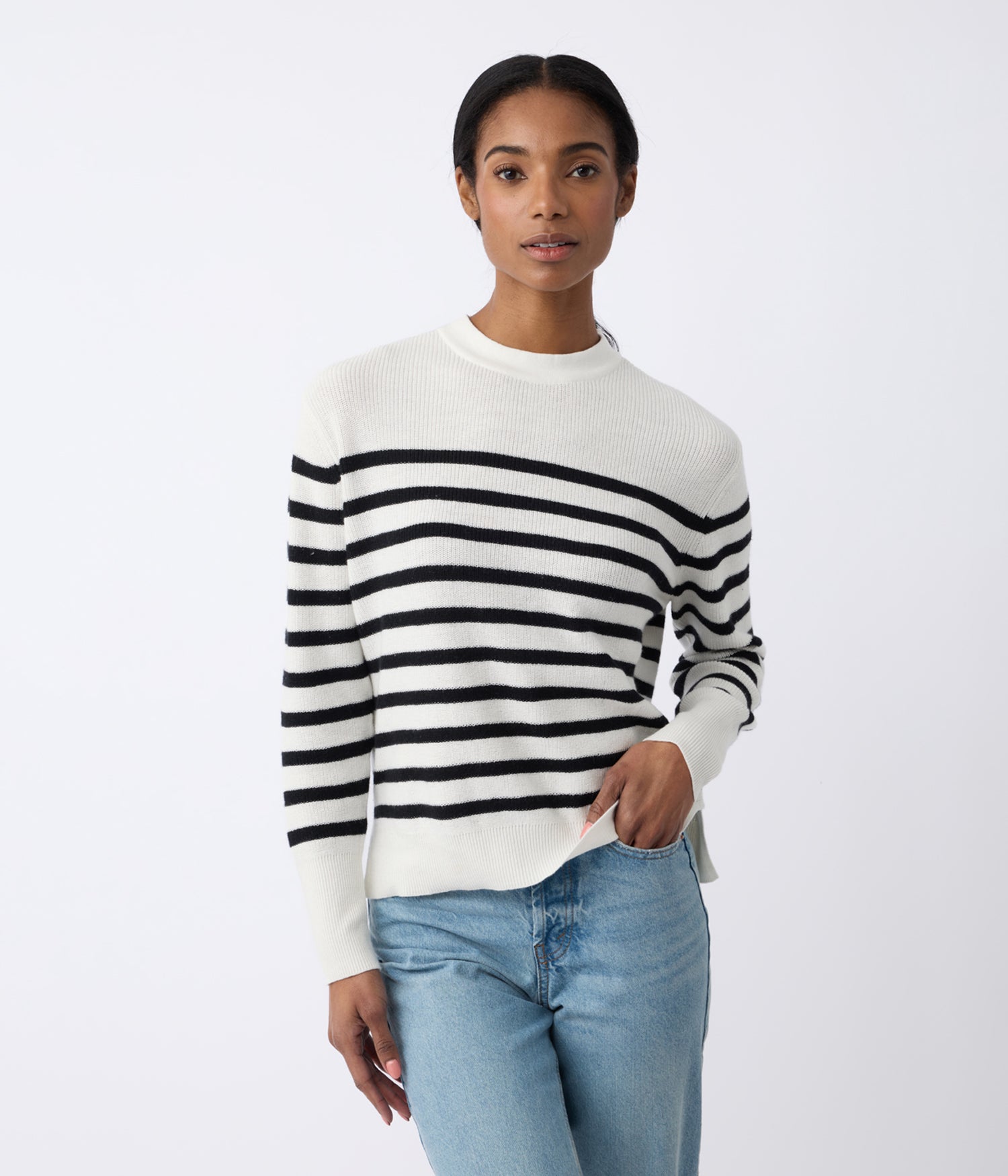 A person in Cozy Earth's white Women's Rowan Sweater with black stripes and light blue jeans stands against a plain white background, exuding calmness with hands casually at their sides. |Color:Bone/Jet Black Stripe