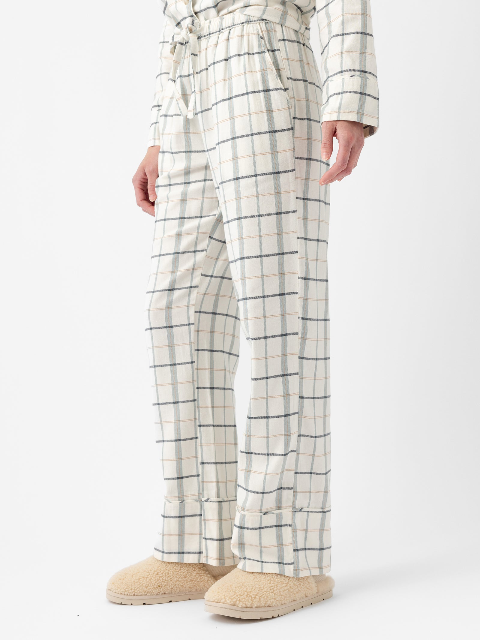 A person is wearing the cream-colored pajama pants from the Cozy Earth Women's Farmhouse Long Sleeve Pajama Set, featuring a blue and gray plaid pattern and a drawstring waist. They are also donning beige, fuzzy slippers, with only their lower half visible against a white background. 