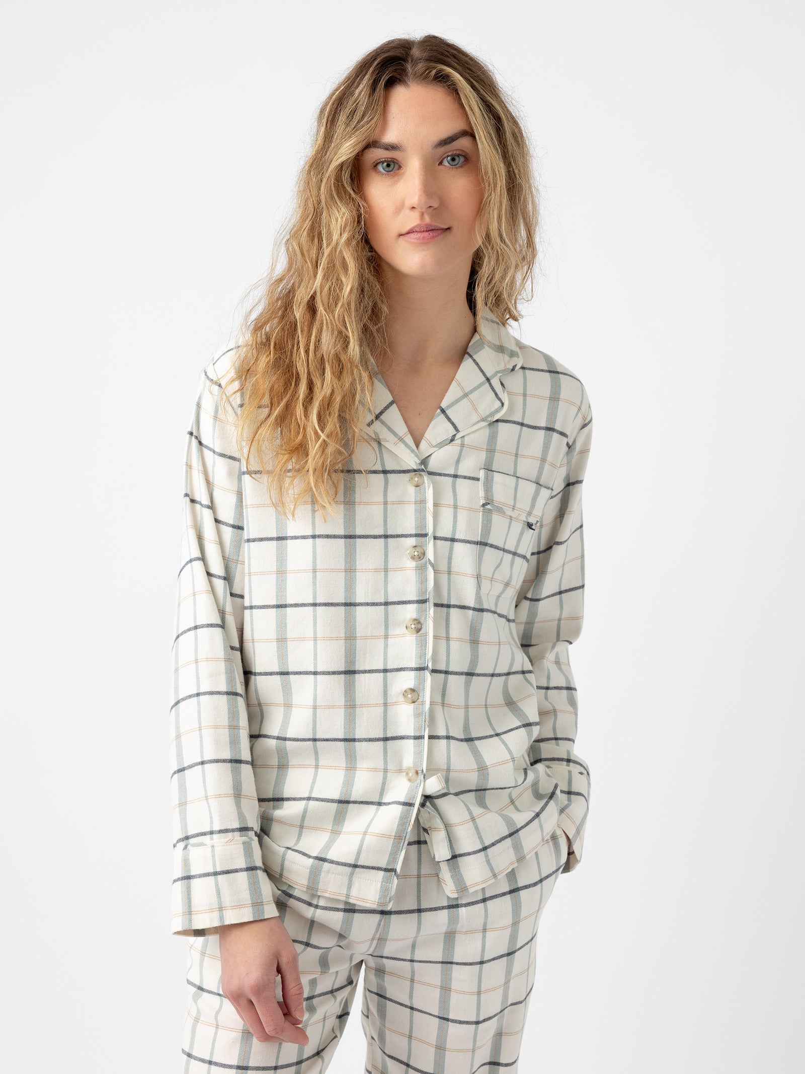 A person with long wavy hair stands against a white background, wearing a matching pajamas set from Cozy Earth. The Women's Farmhouse Long Sleeve Pajama Top features a button-up design with a collar and chest pocket, along with loosely fitting pants, all in a white base and light blue/green plaid pattern. 