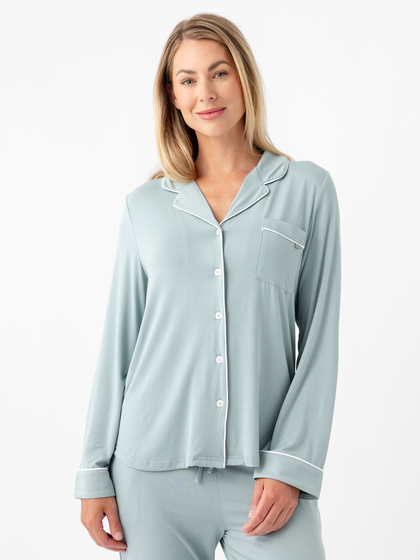 A person with long blonde hair is standing and wearing the HIDE Women's Long Sleeve Bamboo Pajama Top in Stretch-Knit, featuring light blue fabric with white piping. The top includes a collar, buttons down the front, and a pocket on the left side. The person is smiling slightly and has a relaxed posture against a plain white background. 