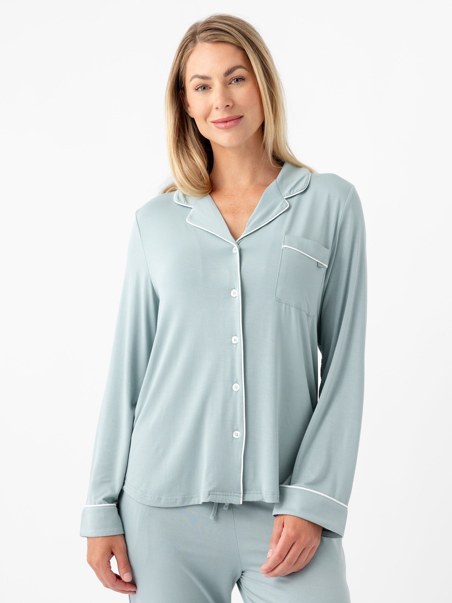 A person with long blonde hair is standing and wearing the HIDE Women's Long Sleeve Bamboo Pajama Top in Stretch-Knit, featuring light blue fabric with white piping. The top includes a collar, buttons down the front, and a pocket on the left side. The person is smiling slightly and has a relaxed posture against a plain white background. |Color:Brook