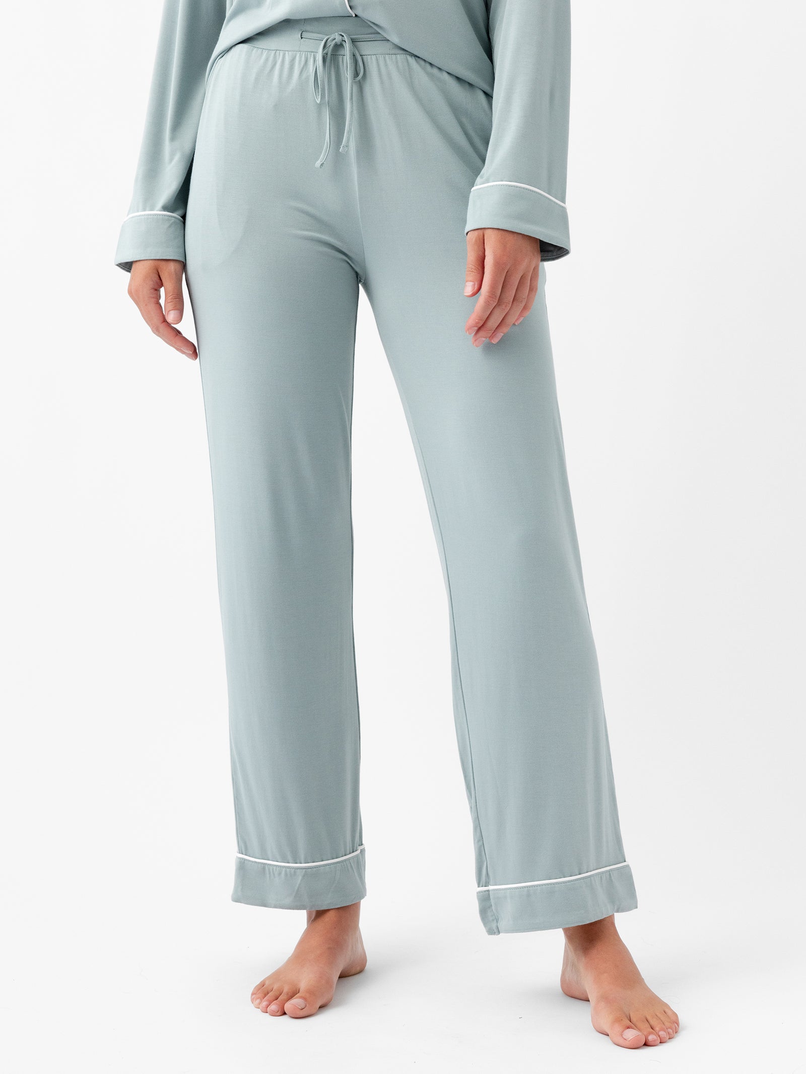 A person stands barefoot wearing Cozy Earth Women's Stretch-Knit Bamboo Pajama Pants in light green with white piping detail. The pajamas feature a relaxed-fit long-sleeved top and drawstring pants, perfect for sleep or lounging. The background is plain white. 