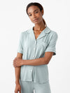 A person with long hair stands against a white background, wearing a pastel green Women's Short Sleeve Bamboo Pajama Top in Stretch-Knit by Cozy Earth. The top features white piping, short sleeves, a collar, buttons down the front, and a chest pocket. The shorts are relaxed in fit. They are smiling softly with arms crossed. |Color:Brook