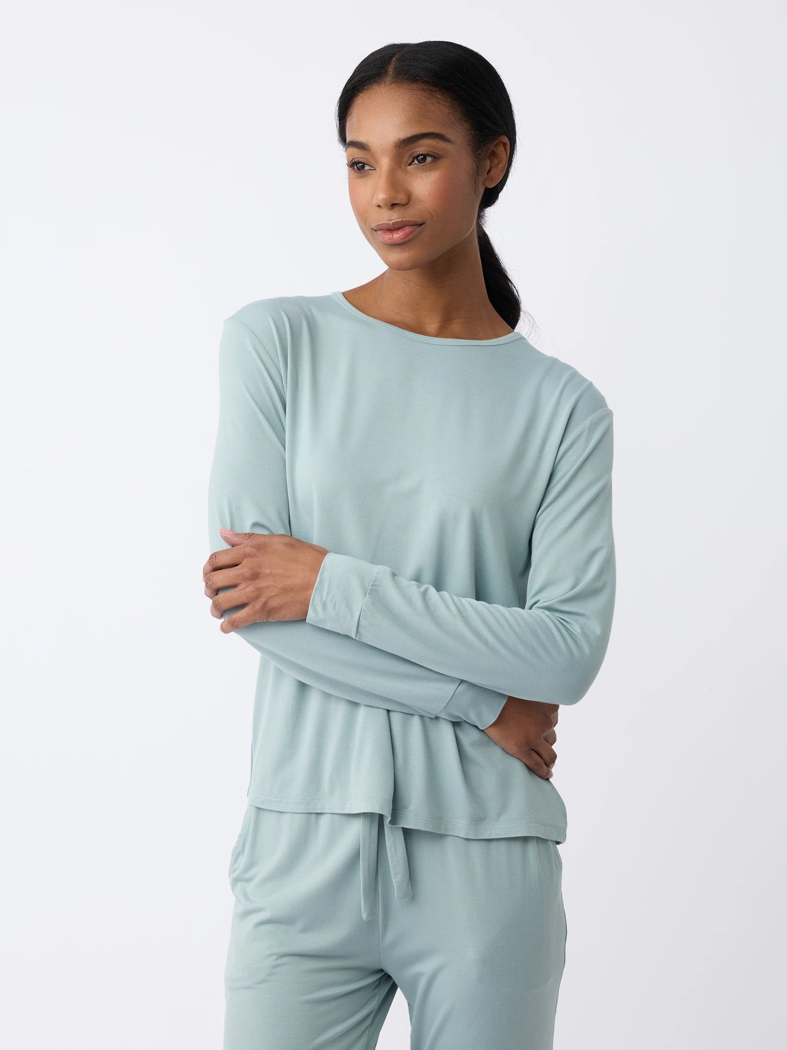 A woman with long hair wears a light blue Women's Stretch Knit Bamboo Long Sleeve Lounge Tee by Cozy Earth and matching pants, arms crossed against a plain white backdrop, gazing sideways with a calm expression. |Color:Brook