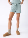 A person wearing Cozy Earth's Stretch Knit Bamboo Shorts in light blue with a drawstring, paired with a matching top. They have on beige socks and fluffy brown slippers against a plain white background. |Color:Brook