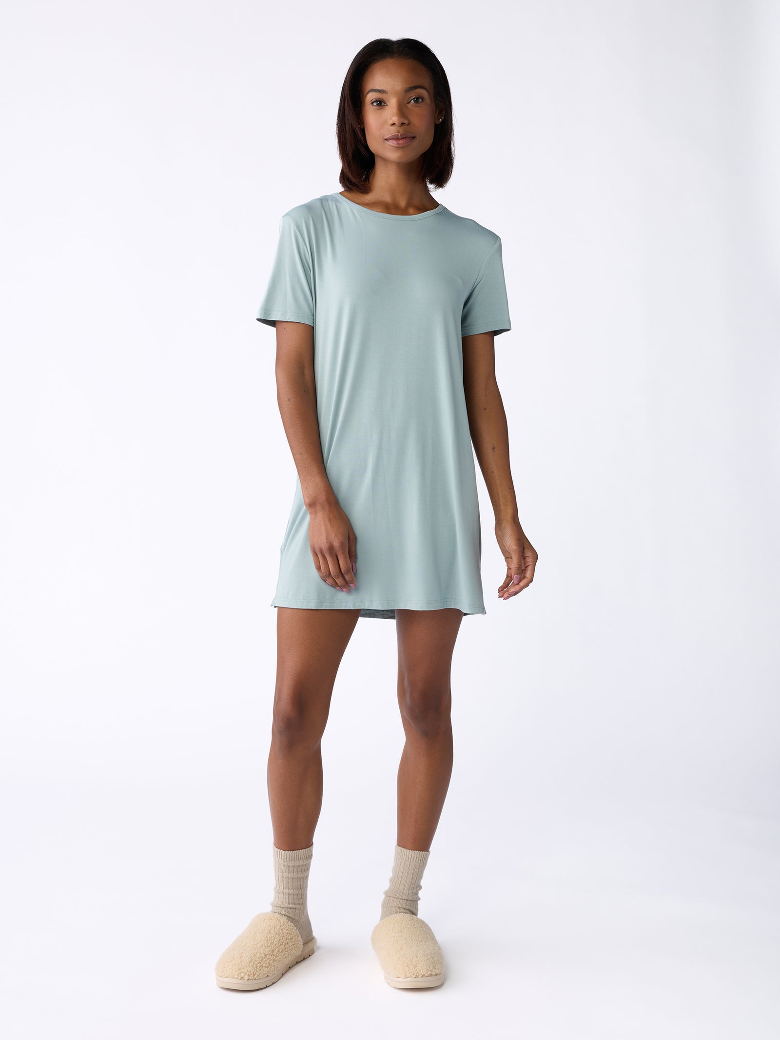 A person wearing the Women's Bamboo Stretch Knit Sleep Dress by Cozy Earth in light blue, along with beige socks and cozy beige slippers, stands against a plain white background. 