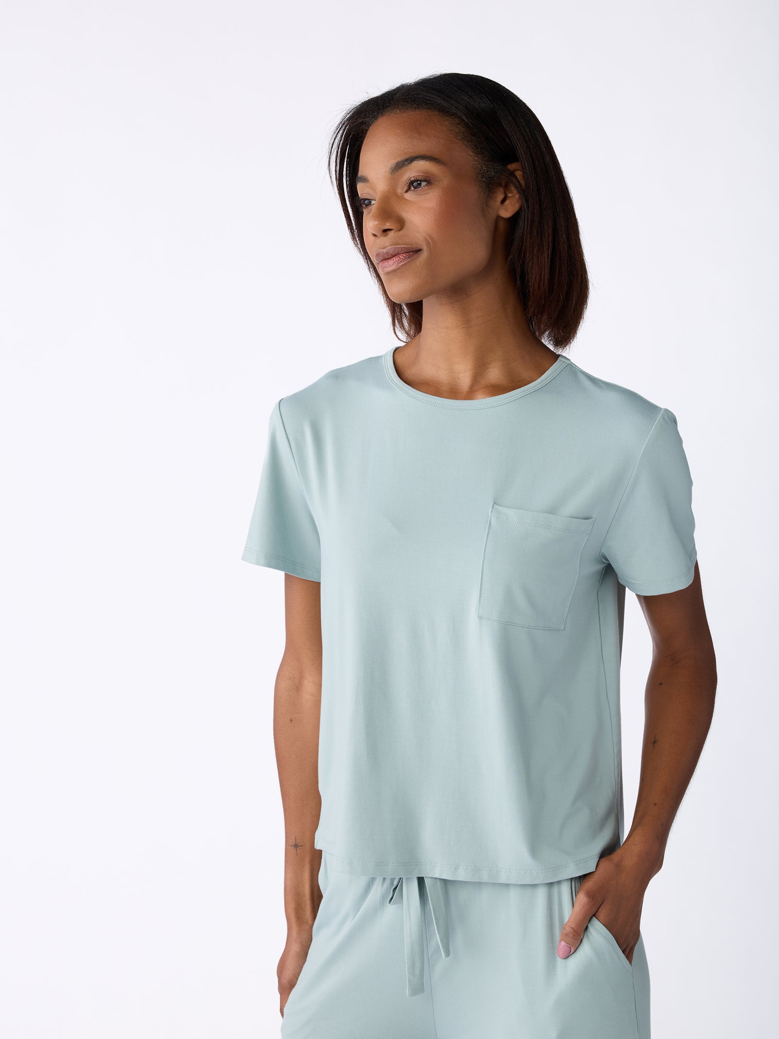 A person wearing a Cozy Earth Women's Stretch-Knit Bamboo Lounge Tee in light blue stands against a plain white background. The short-sleeved shirt features a pocket, and the individual has their right hand in the pocket of matching pants while looking slightly away from the camera. 