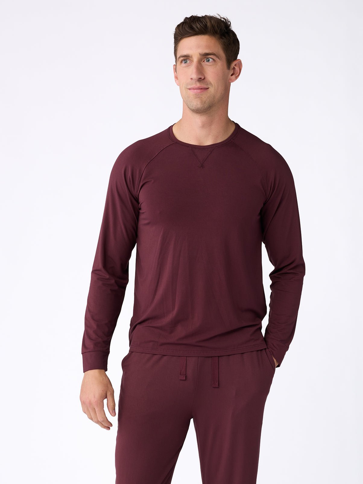 A person dressed in a Cozy Earth Men's Stretch-Knit Bamboo Long Sleeve in burgundy, along with matching pants, stands against a plain white background, gazing slightly to the side. 