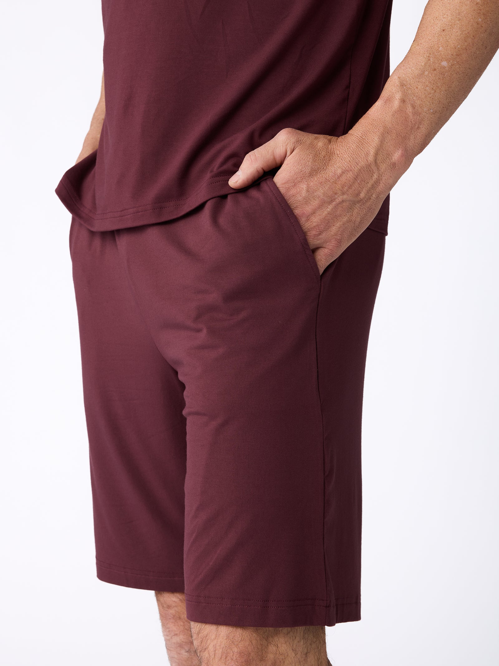 A person dressed in an outfit featuring the Men's Stretch-Knit Bamboo Pajama Short from Cozy Earth, paired with a burgundy short-sleeved shirt, stands with one hand in their pocket against a plain white backdrop. 