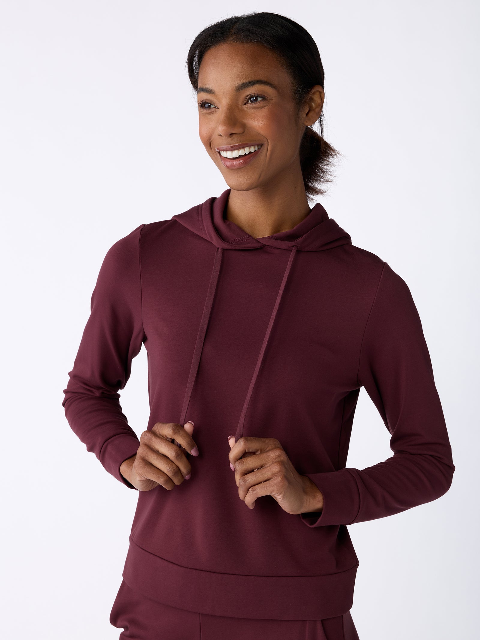 A person wearing a Cozy Earth Women's Ultra-Soft Bamboo Hoodie in maroon smiles while facing slightly to the right, with their hands holding the hoodie’s drawstrings. The background is a simple, light gray color. 