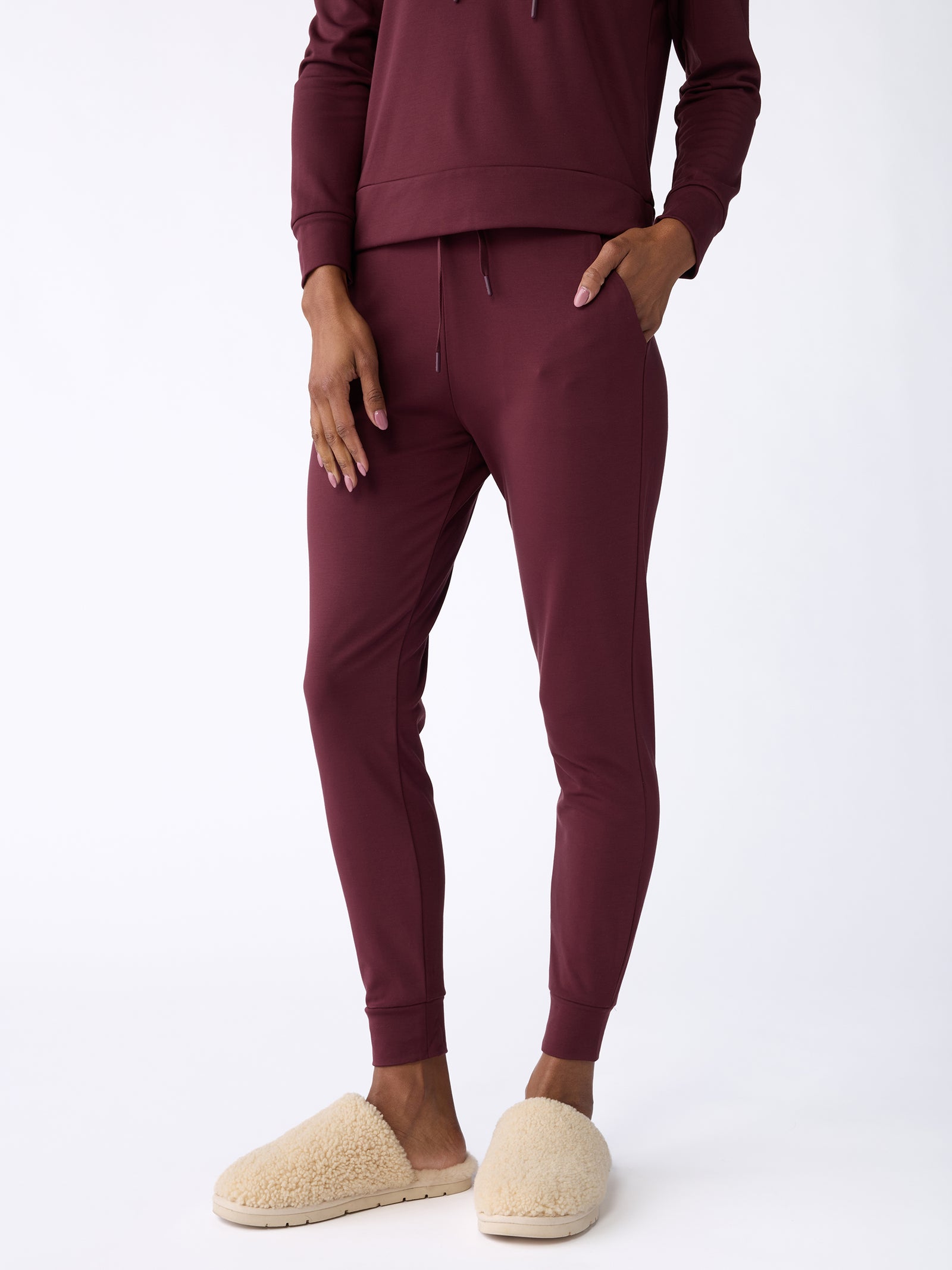 A person is wearing Cozy Earth's Women's Bamboo Jogger Pants in burgundy with their hands in the pockets, paired with beige fuzzy slippers, standing against a plain white background. 