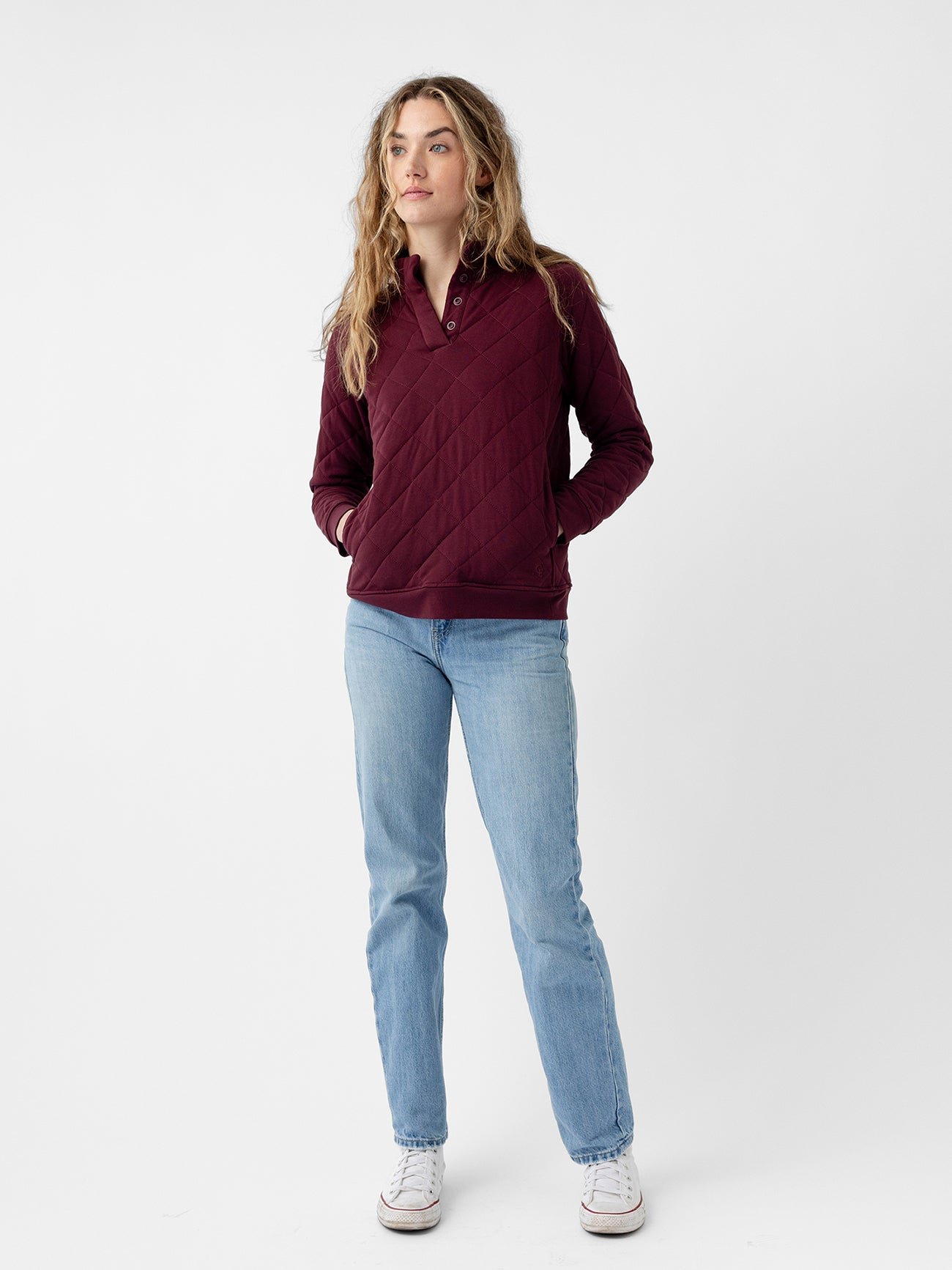 A person with long, wavy hair stands against a plain white background, wearing the Women's Ultra-Soft Bamboo Quilted Snap Pullover by Cozy Earth in maroon, light blue jeans, and white sneakers. Their hands are in their pockets, and they are looking slightly to the side. 