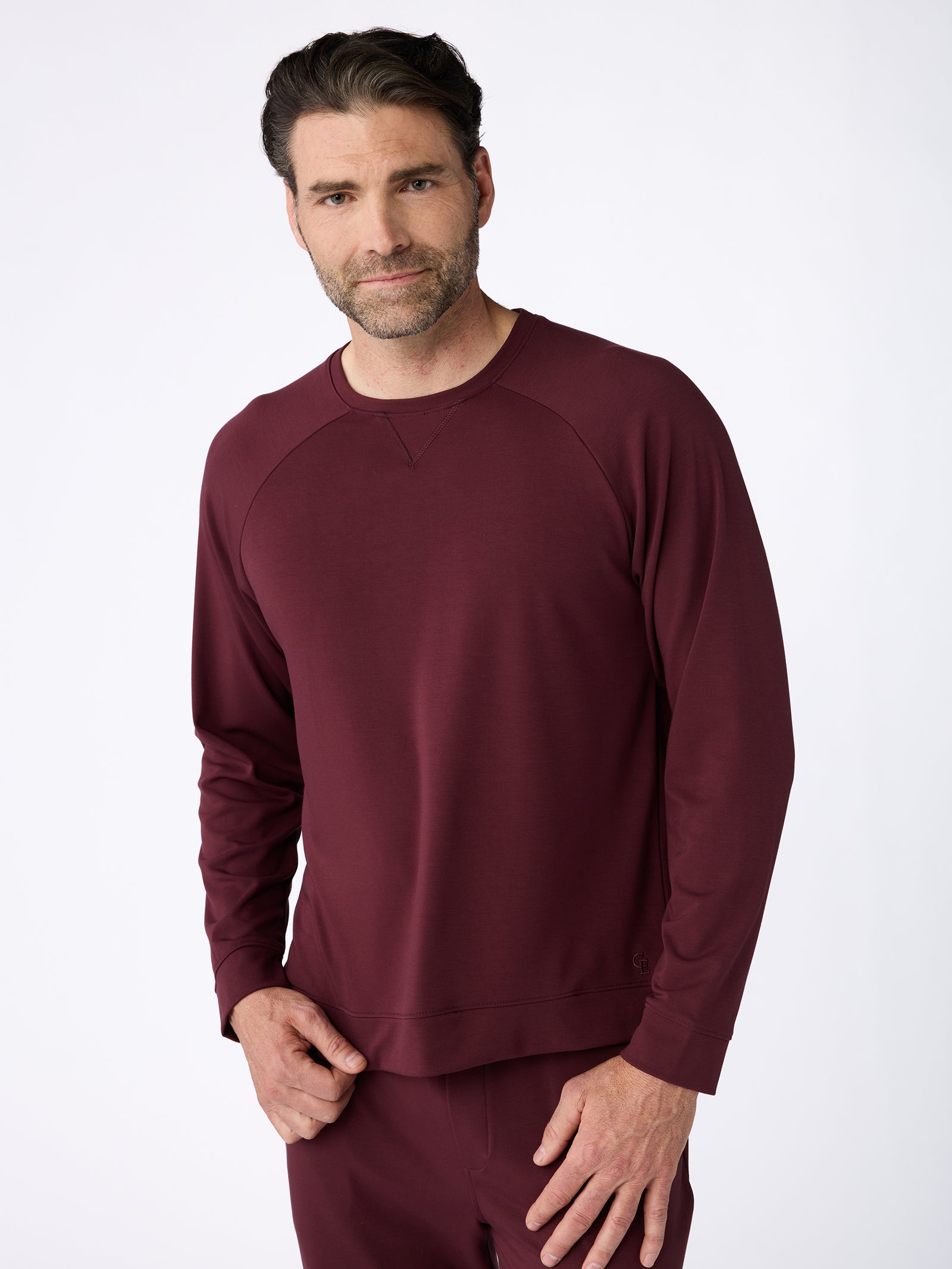 A man with short dark hair and facial hair is wearing a maroon Men's Ultra-Soft Bamboo Pullover Crew by Cozy Earth, along with matching pants. He stands against a plain white background, looking towards the camera with a small smile. 