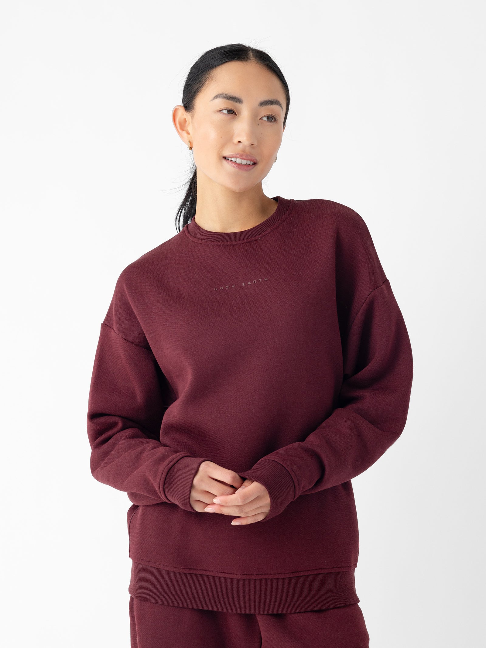 A person with dark hair tied back is wearing the Cozy Earth Women's CityScape Crewneck, paired with matching pants. They are standing against a plain white background. The crewneck features subtle lettering on the front. The person is smiling gently and has their hands clasped together. 