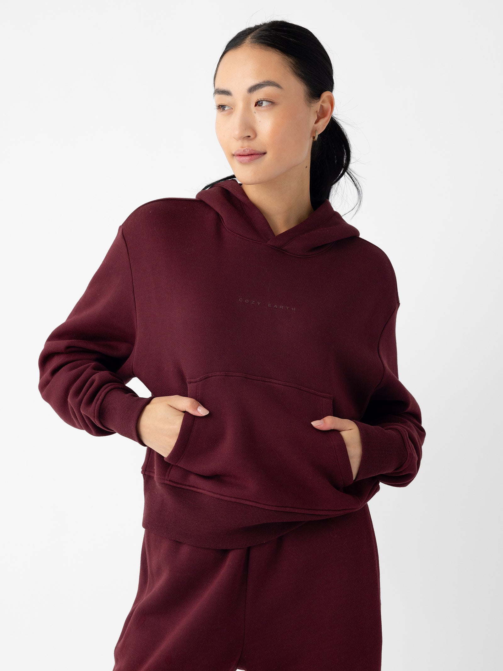 A person with long dark hair tied back is wearing a Women's CityScape Cropped Hoodie and matching sweatpants from Cozy Earth in a maroon shade. They have their hands in the hoodie pockets and are looking slightly to the side. The background is plain white. 