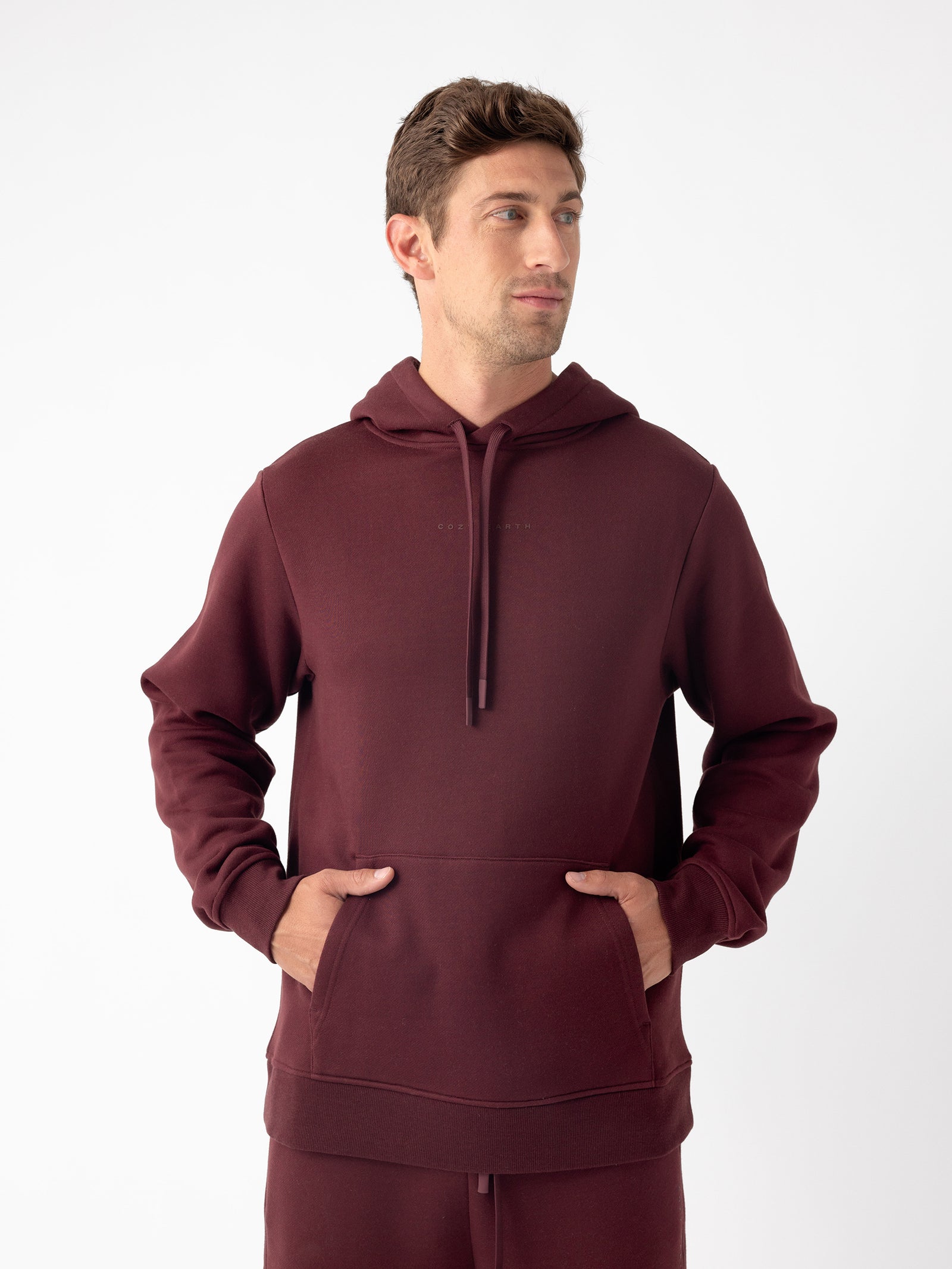 A man is wearing a maroon Men's CityScape Hoodie by Cozy Earth, looking slightly to the left. He has short brown hair and his hands are tucked into the front pocket of the hoodie. The background is plain white. 