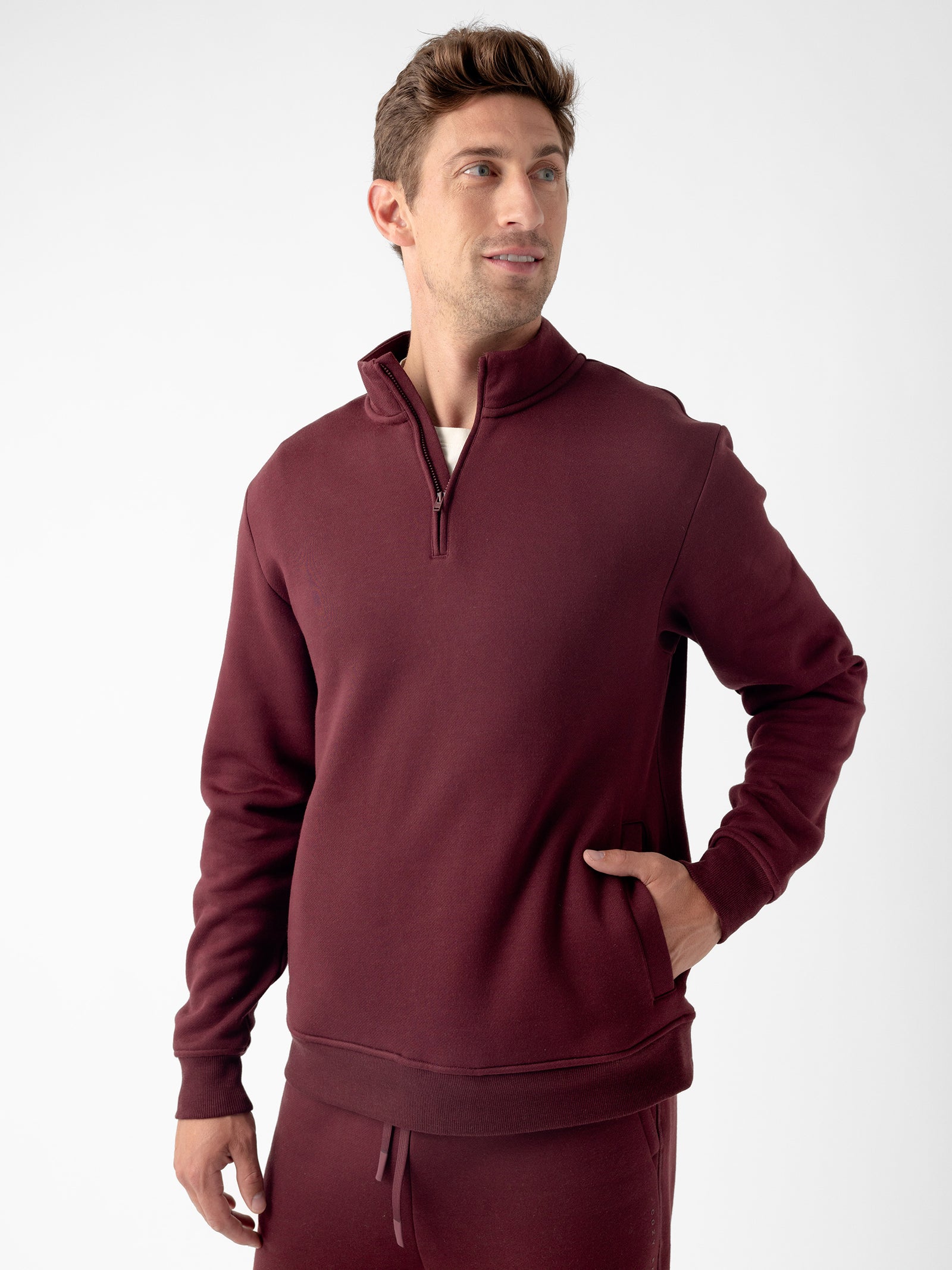 A man stands against a plain background, wearing the Cozy Earth Men's CityScape Quarter Zip in maroon with one hand in his pocket. He has short brown hair and is looking slightly to the side with a relaxed expression. He is also wearing matching maroon sweatpants. 