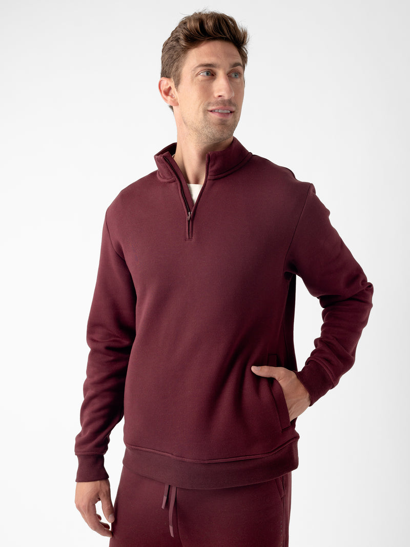 A man stands against a plain background, wearing the Cozy Earth Men's CityScape Quarter Zip in maroon with one hand in his pocket. He has short brown hair and is looking slightly to the side with a relaxed expression. He is also wearing matching maroon sweatpants.