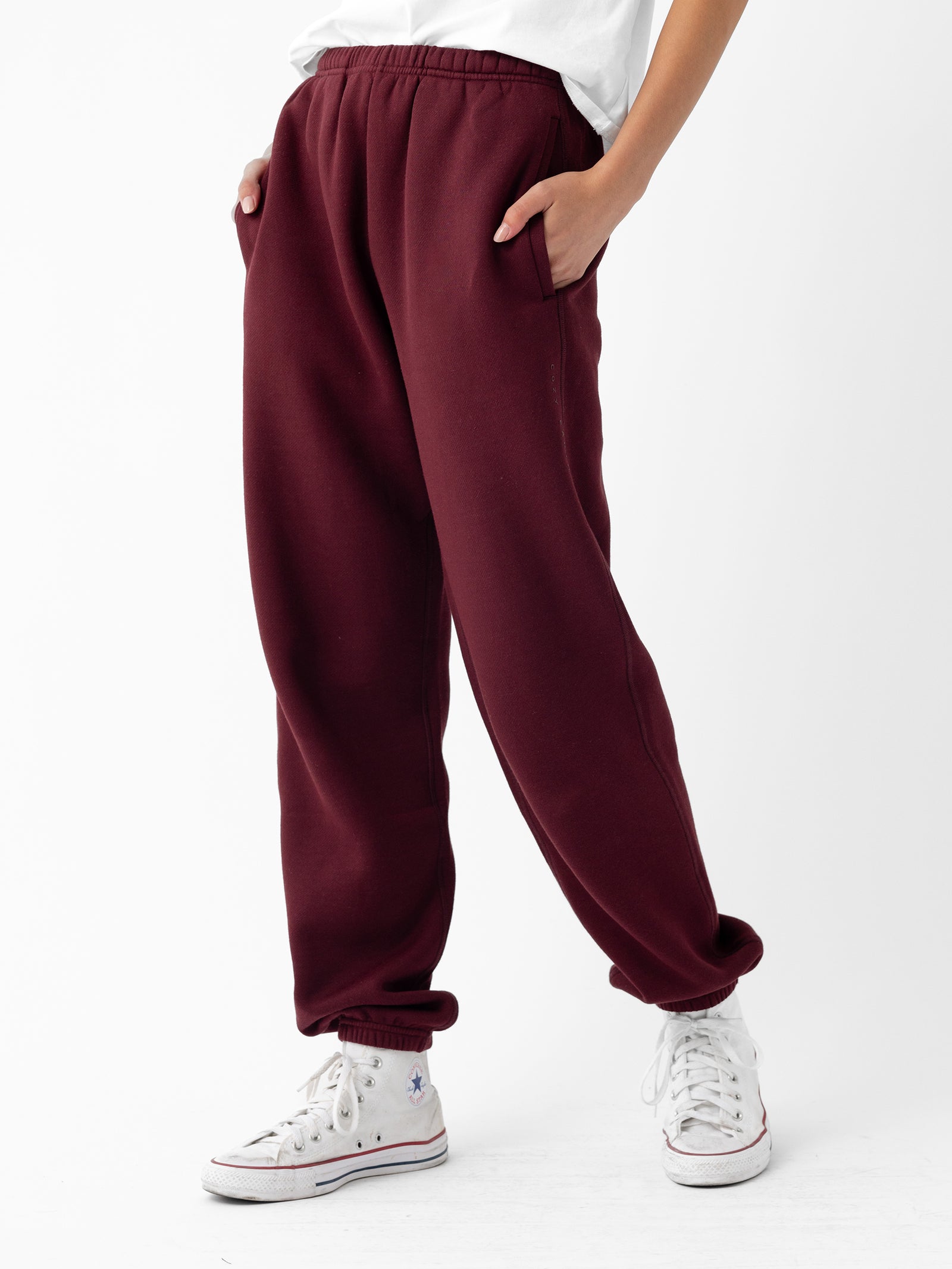 A person's lower body is visible, showcasing the Women's CityScape Sweatpant by Cozy Earth. The maroon sweatpants feature hands in pockets and rolled cuffs, paired with white sneakers, set against a plain white background. 