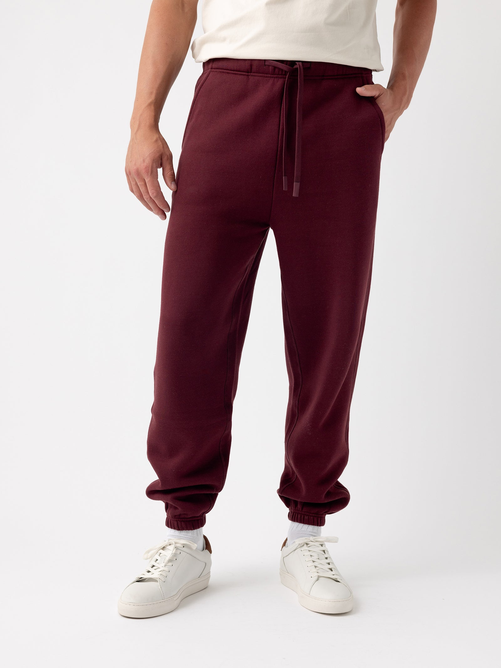 A person in a cream-colored shirt is wearing maroon Men's CityScape Sweatpant by Cozy Earth with drawstrings and elastic cuffs at the ankles, paired with white sneakers. The person has one hand in a pocket and is standing against a plain, light background. 