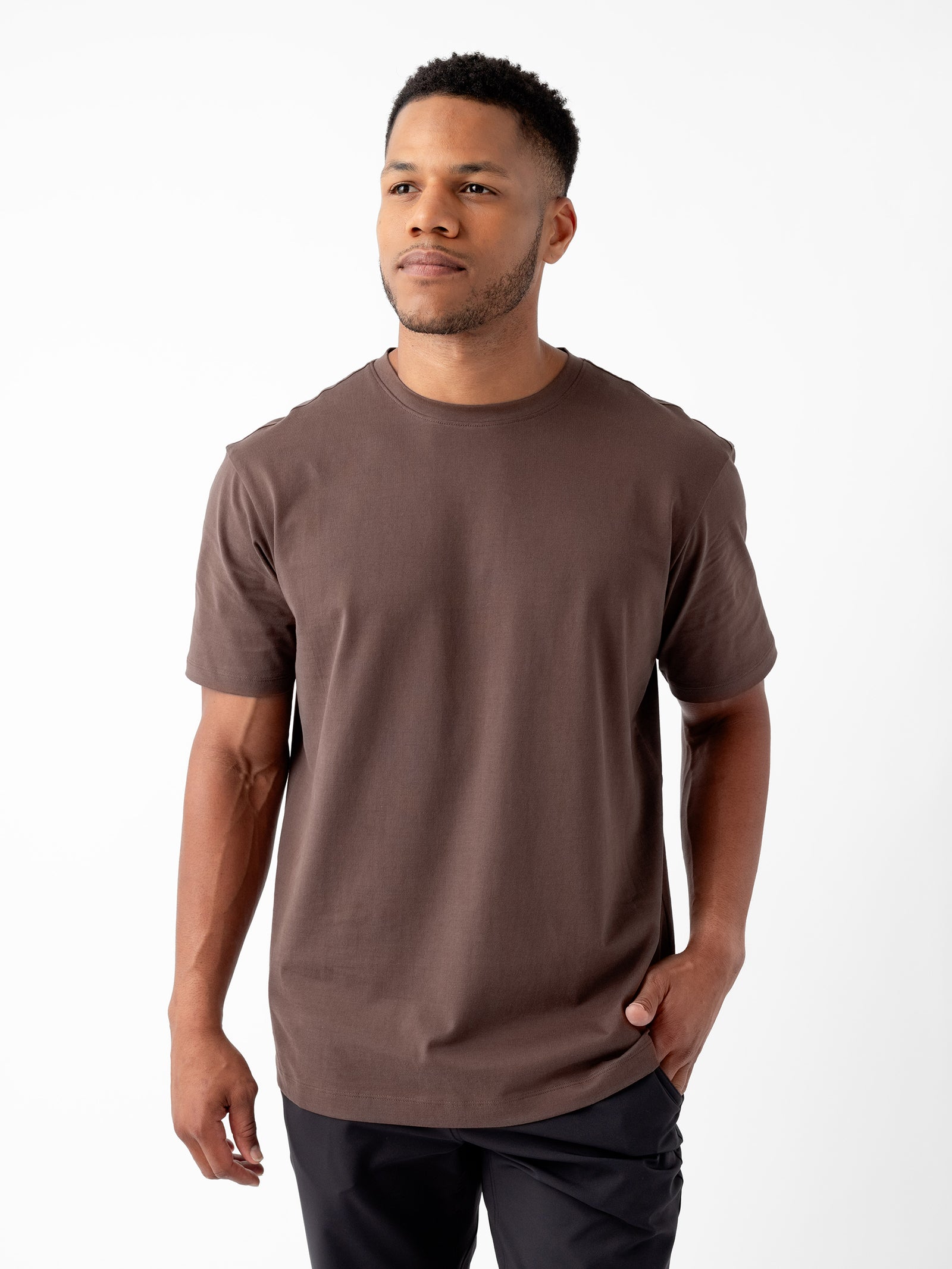 A man with short black hair and a short beard is wearing a brown Men's All Day Tee from Cozy Earth along with black pants. He stands facing slightly to the left with a hand in his pocket, against a plain white background. 