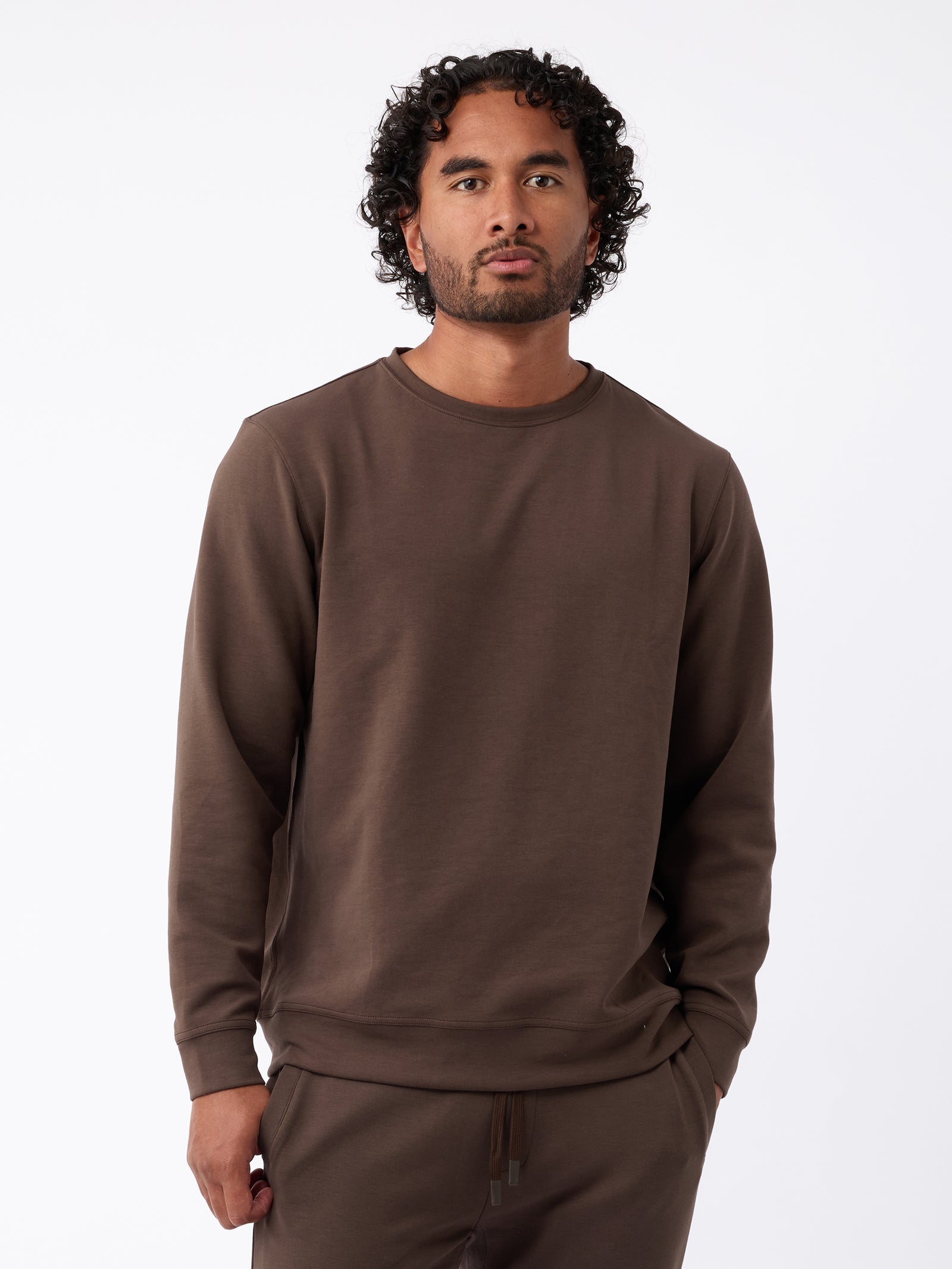 A man with curly hair and a beard wears the Men's StretchTech Crewneck by Cozy Earth in brown, along with matching pants. He stands against a plain white background with his hands in his pockets, looking straight at the camera. 