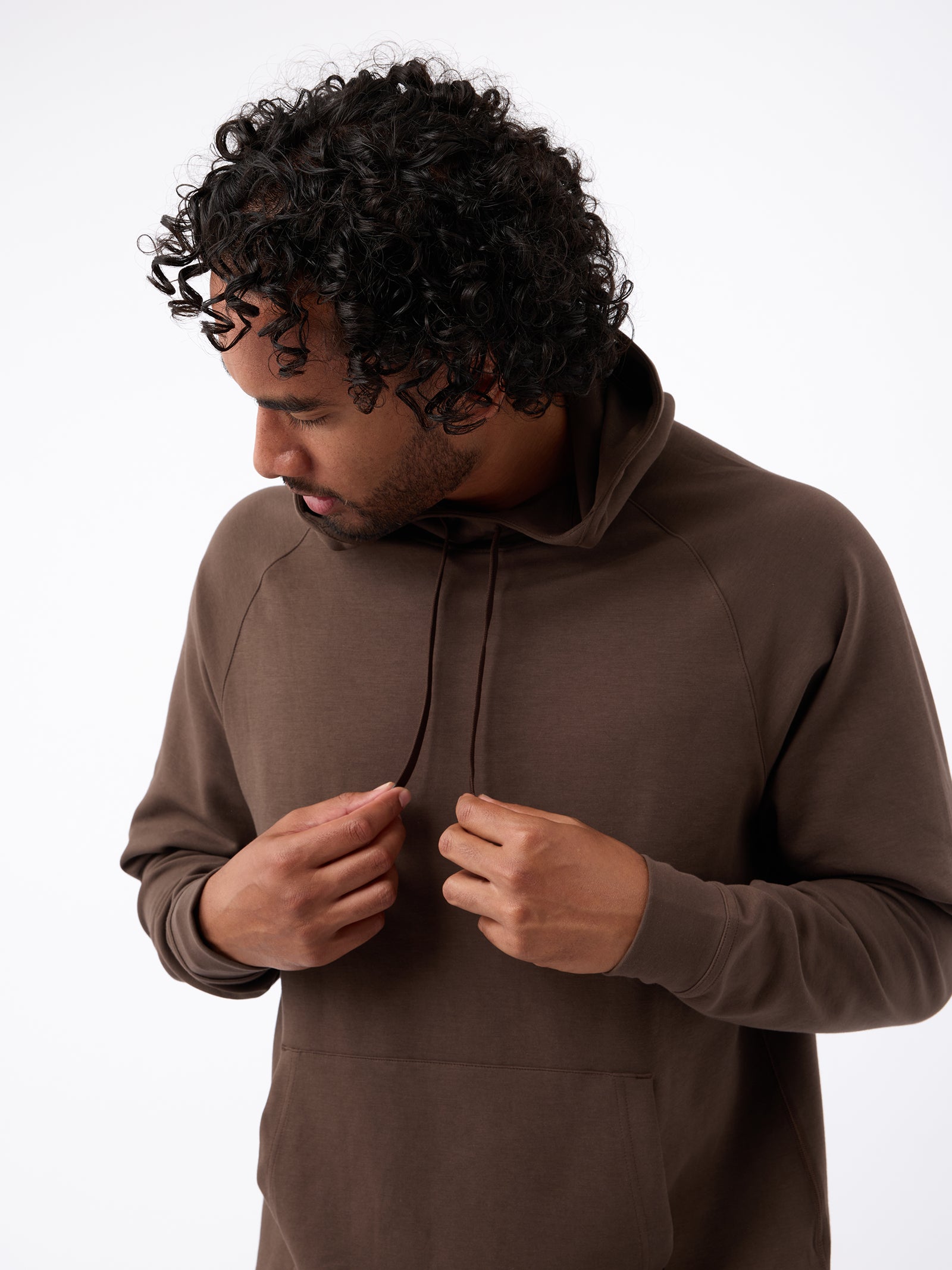 A person with curly hair is looking down and adjusting the drawstrings of their brown Men's StretchTech Hoodie from Cozy Earth. They are wearing a casual outfit and standing against a plain white background. 