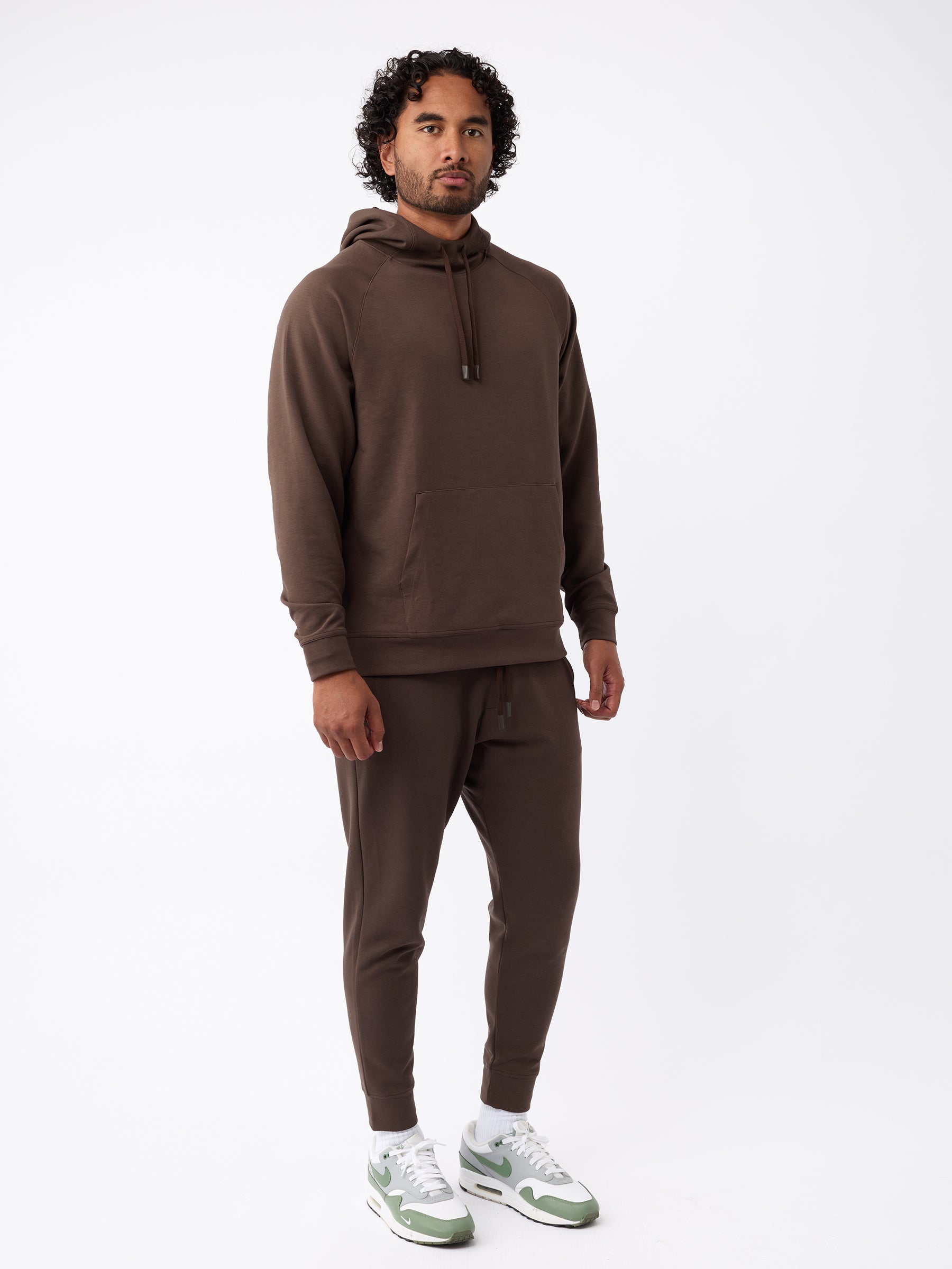 A man stands against a plain white background, wearing Cozy Earth's Men's StretchTech Jogger along with a matching brown hoodie. He has dark curly hair and a beard, and is also wearing white sneakers with green accents. |Color:Cacao