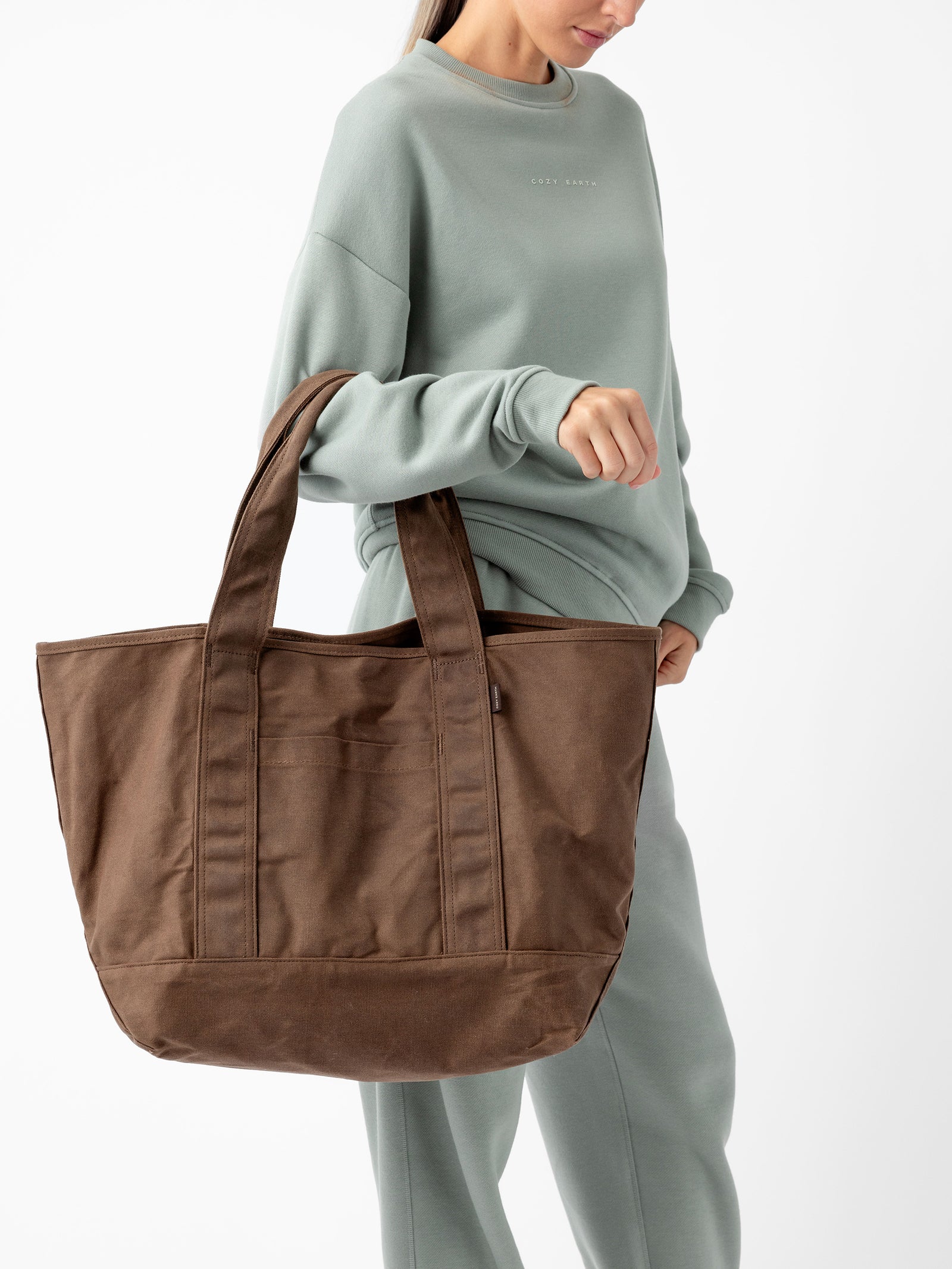 Person wearing a light green sweatshirt and matching pants, holding the Cozy Earth Waxed Canvas Tote by its handles. The background is plain white. 