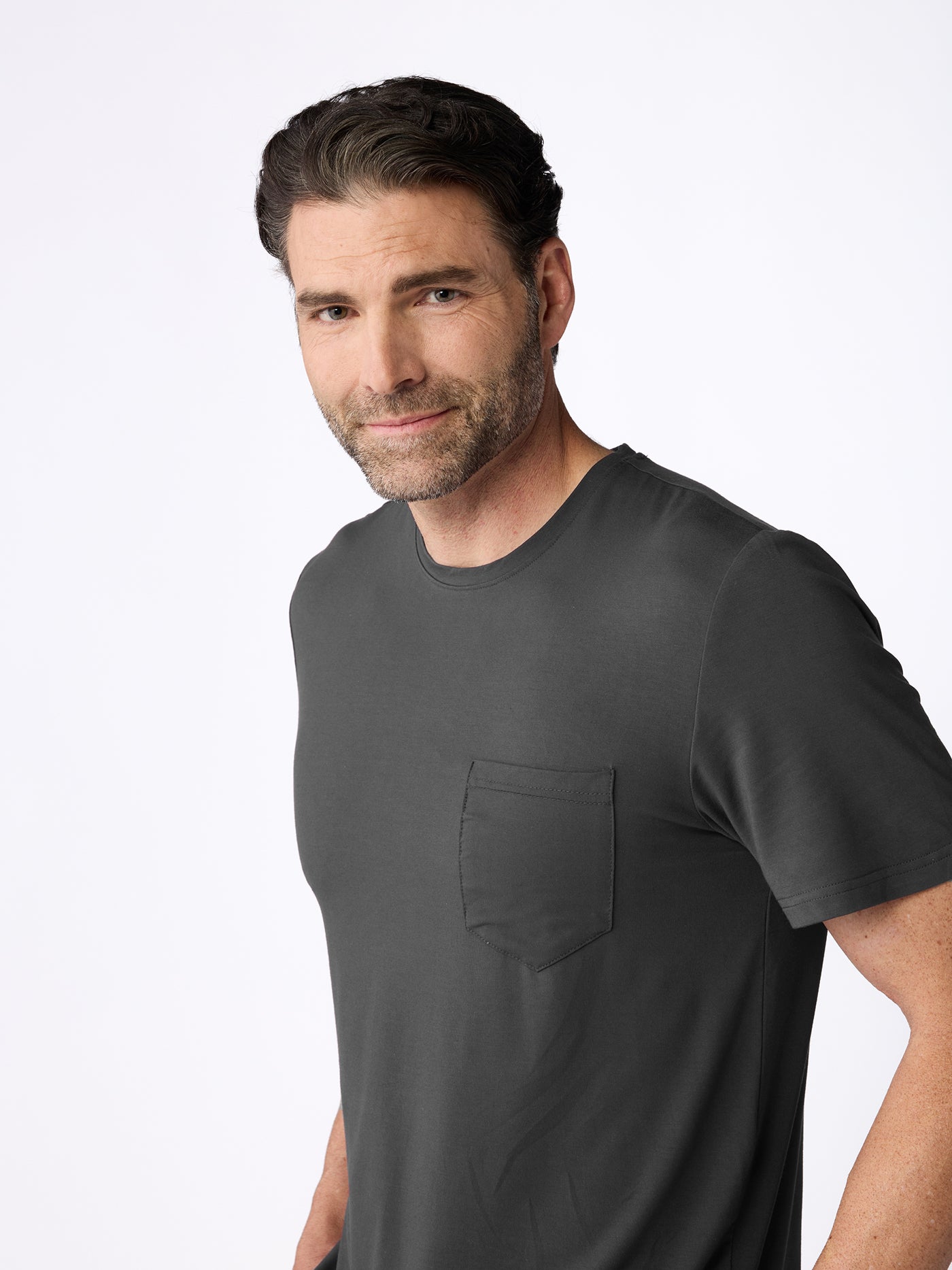 A man with short brown hair and a beard is wearing the Cozy Earth Men's Bamboo Stretch-Knit Short Sleeve Pajama Set in dark gray, standing against a plain white background and smiling slightly at the camera. |Color:Charcoal
