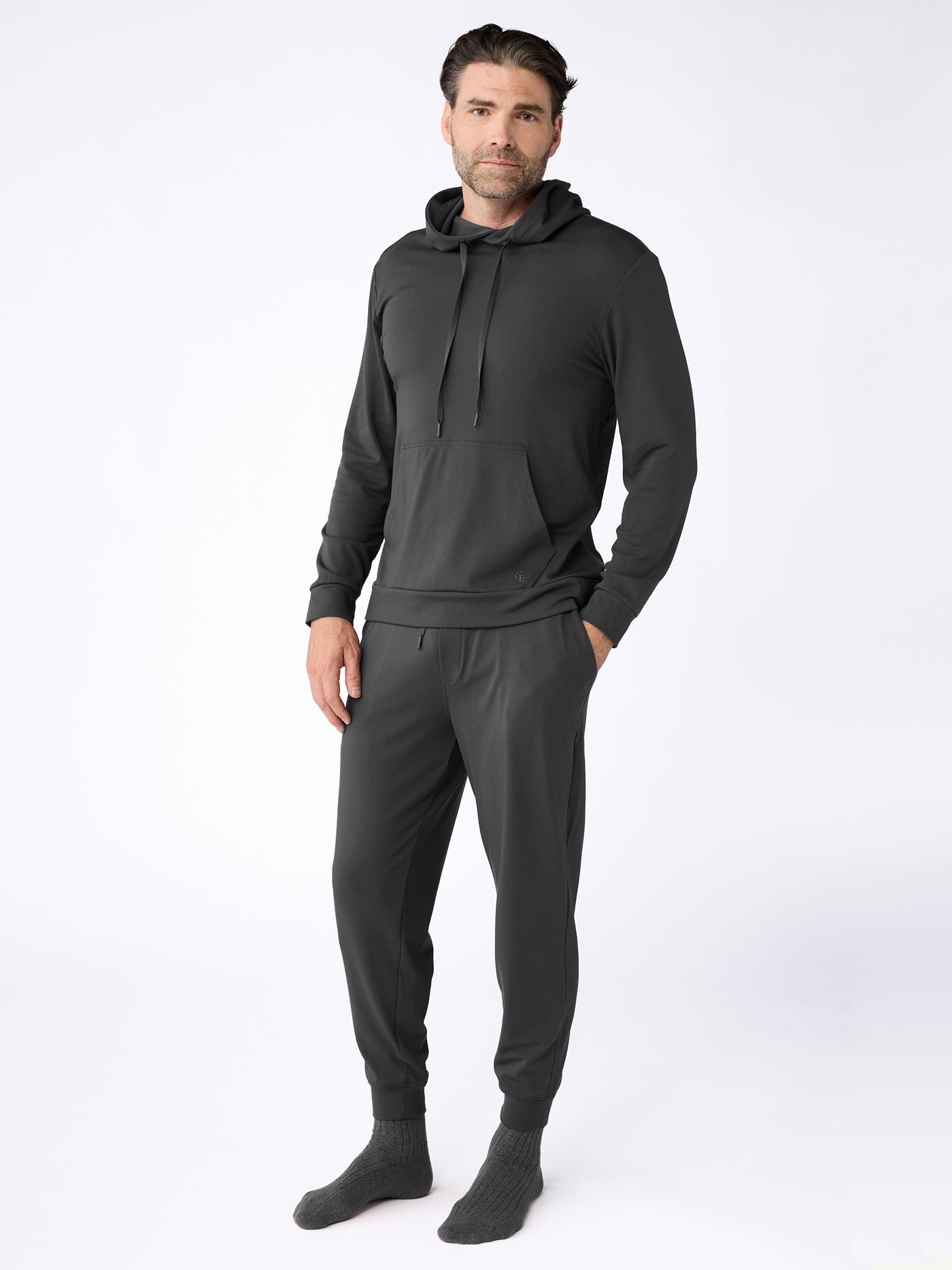 A man wears the Cozy Earth Men's Brushed Bamboo Hoodie & Jogger Set in dark gray, with his hands in his pockets and gray socks, posing against a plain white background. |Color:Charcoal