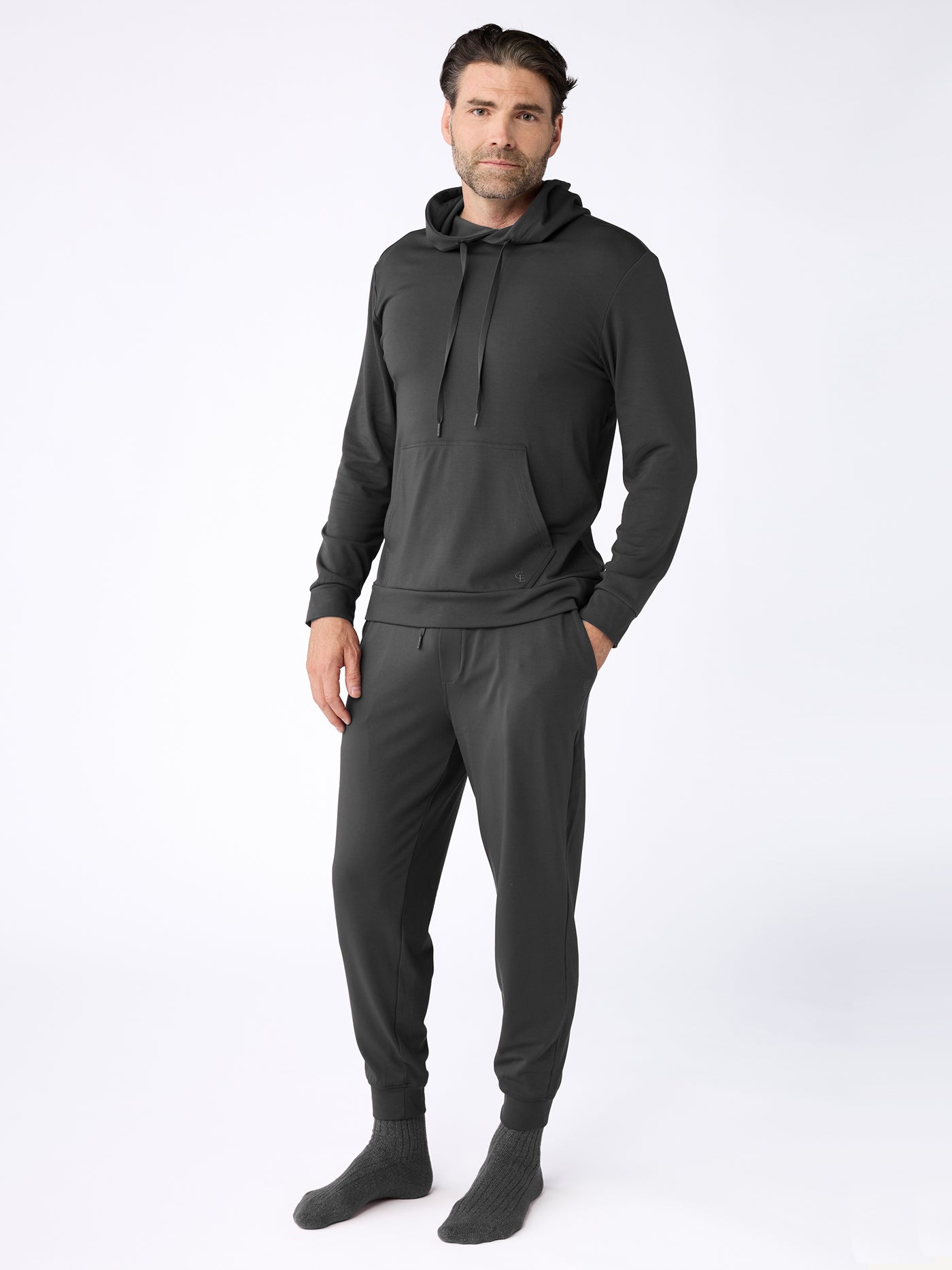 A man wears the Cozy Earth Men's Brushed Bamboo Hoodie & Jogger Set in dark gray, with his hands in his pockets and gray socks, posing against a plain white background. 
