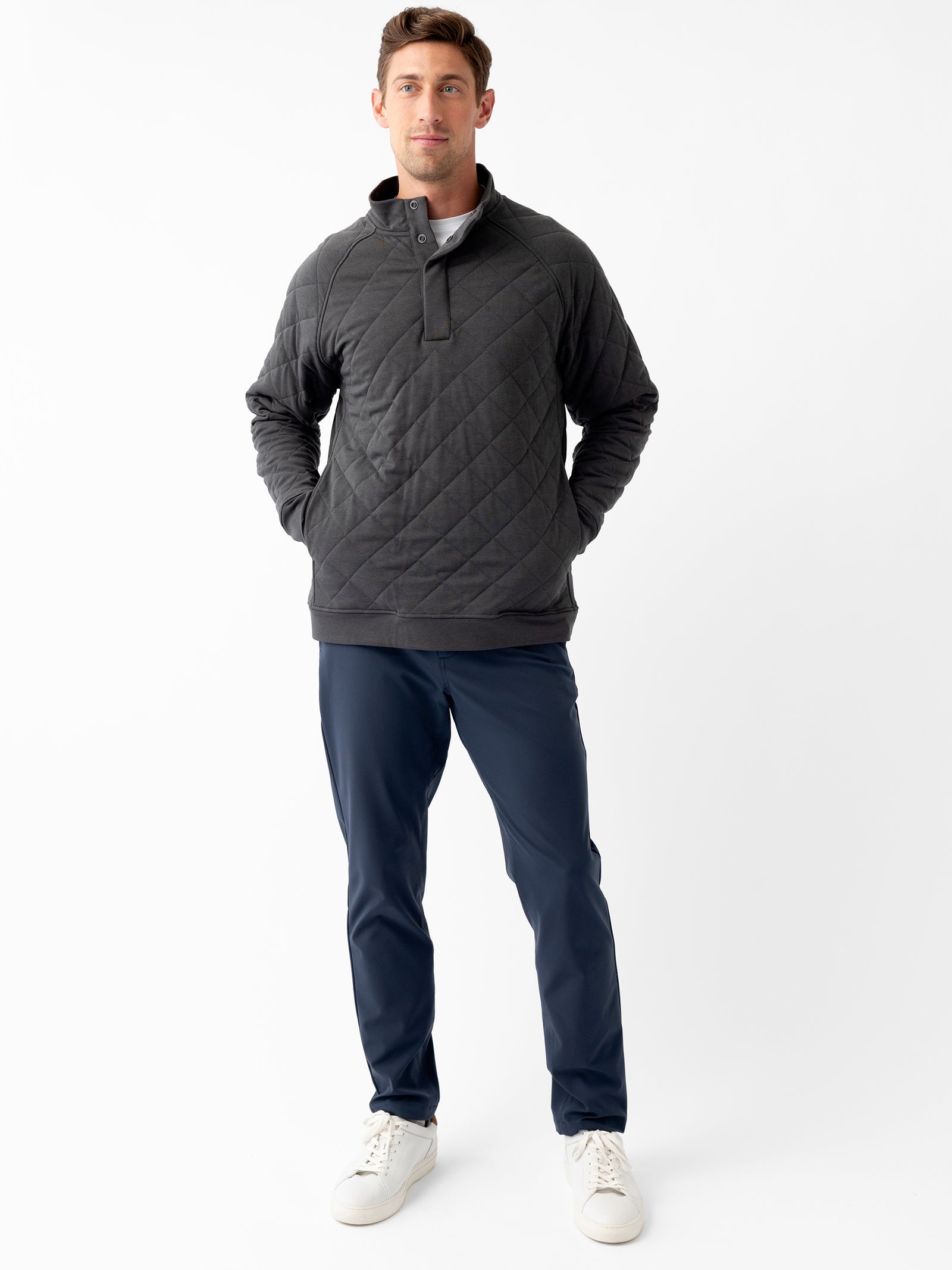 A man stands against a plain white background, wearing the Men's Ultra-Soft Bamboo Quilted Snap Pullover in dark grey by Cozy Earth and navy blue pants. He has his hands in his pockets and is also wearing white sneakers. He is looking at the camera with a neutral expression. 