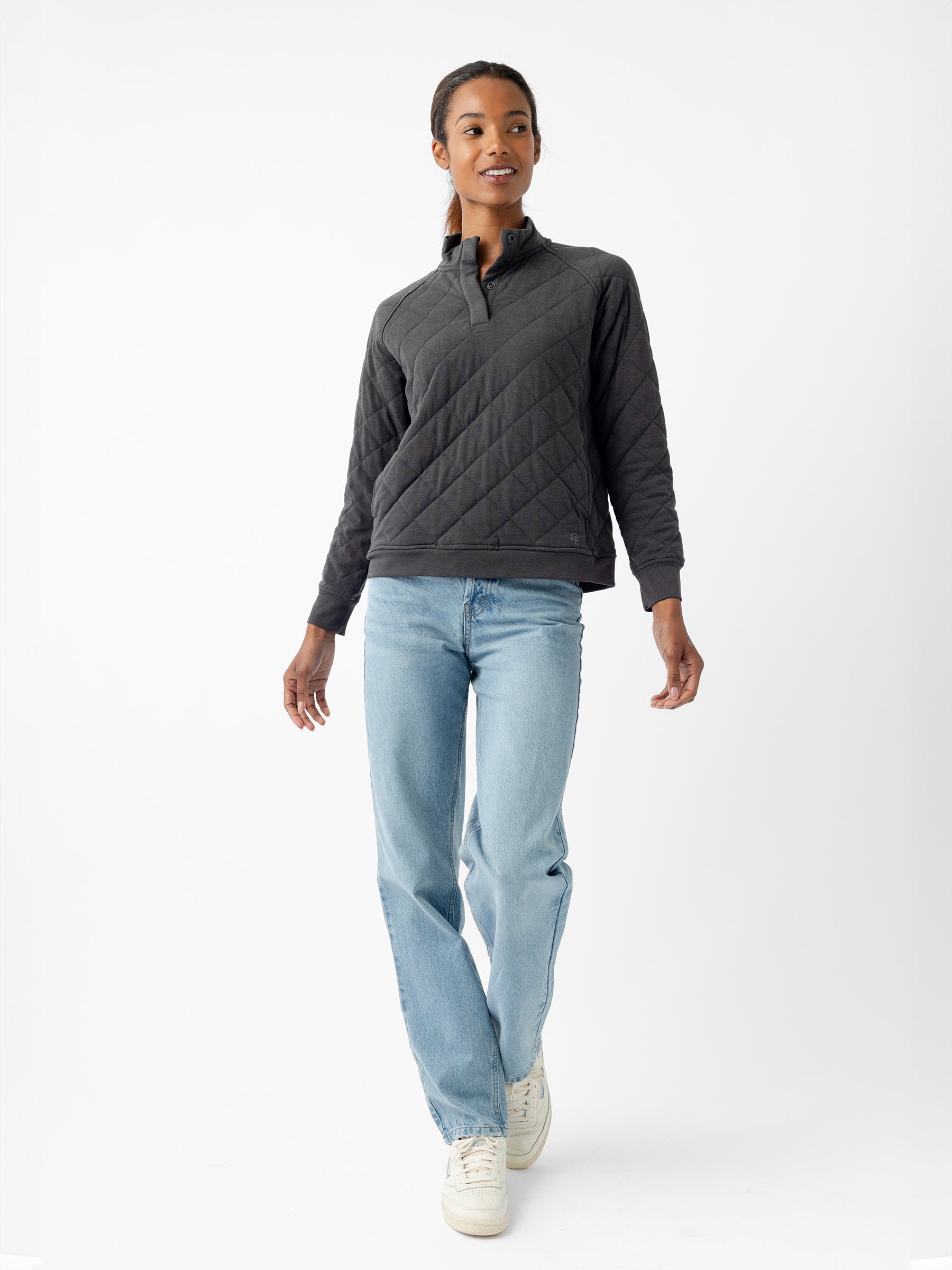 A person with long dark hair is walking while looking slightly to the right. They are wearing a Women's Ultra-Soft Bamboo Quilted Snap Pullover by Cozy Earth, light blue jeans, and white sneakers. The background is a plain white studio setting. |Color:Charcoal