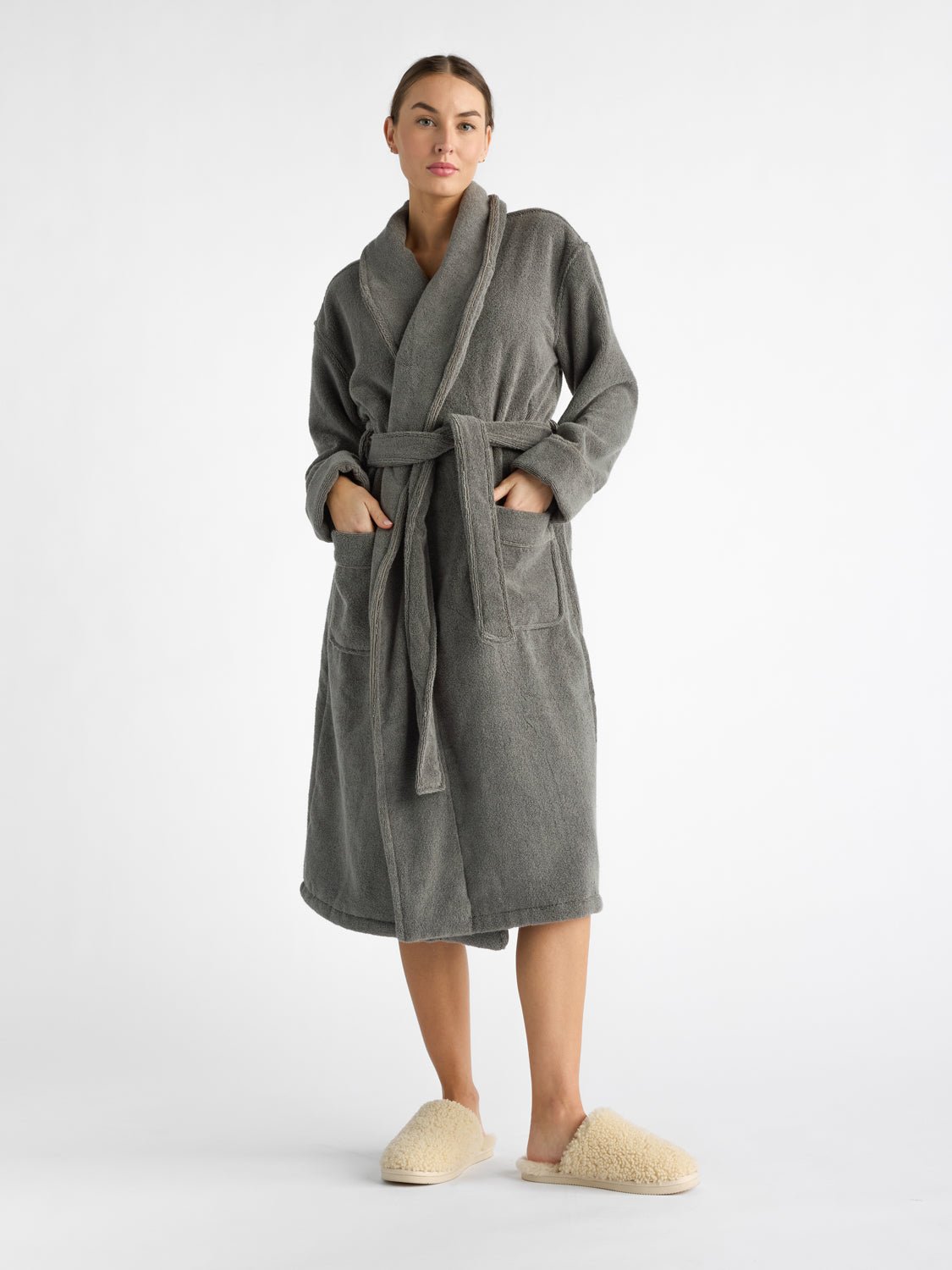 A person models the Luxe Bath Robe by Cozy Earth, featuring charcoal fabric. They stand in slippers against a white background, gazing forward and resting their hands in the pockets of the robe |Color:Charcoal