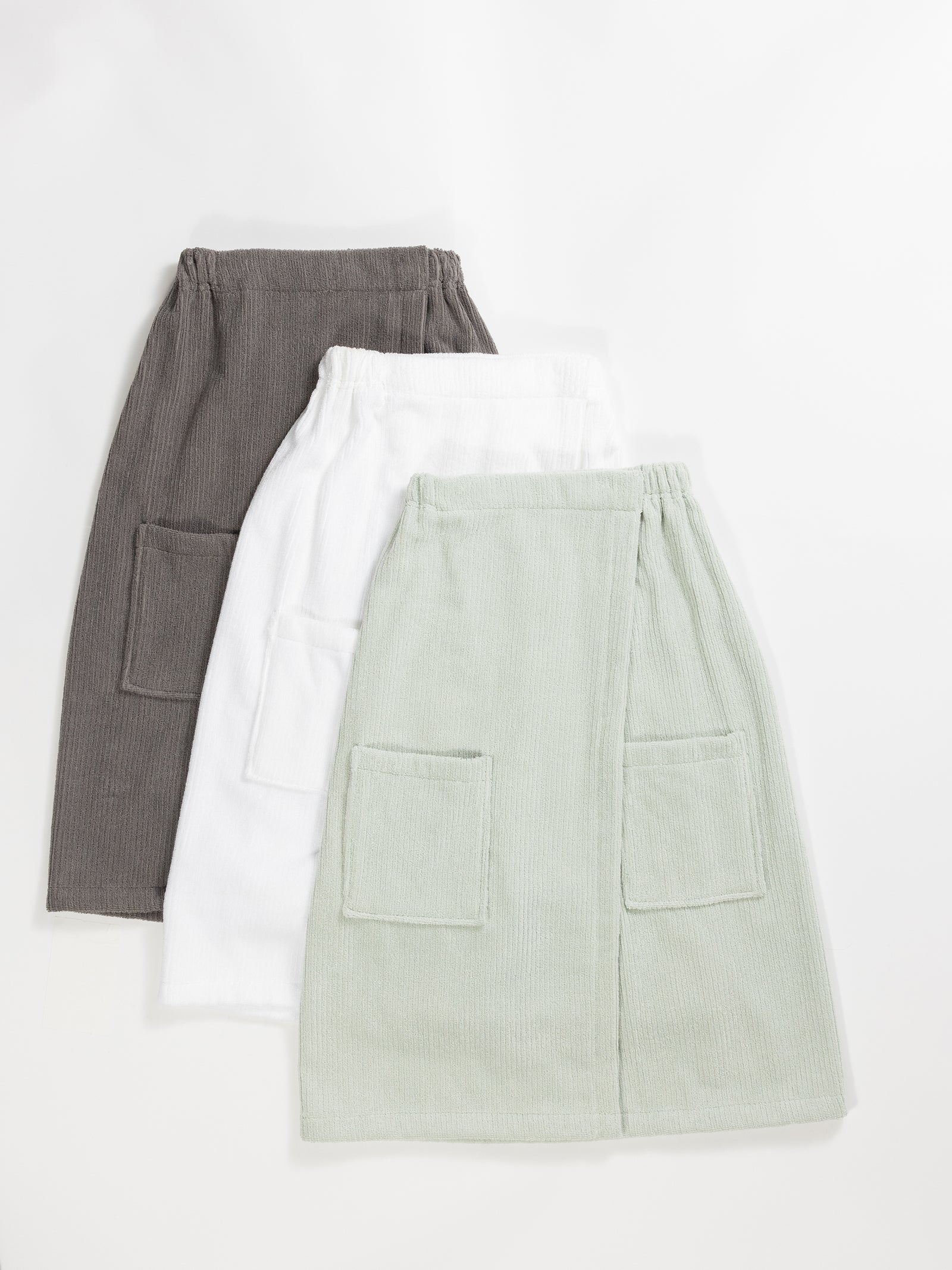 Three Ribbed Terry Bath Wraps by Cozy Earth are arranged in a staggered formation. From left to right, the wraps are dark grey, white, and pale green. Each wrap features an elastic waistband and two front pockets. The fabric appears to be a soft, textured material. 