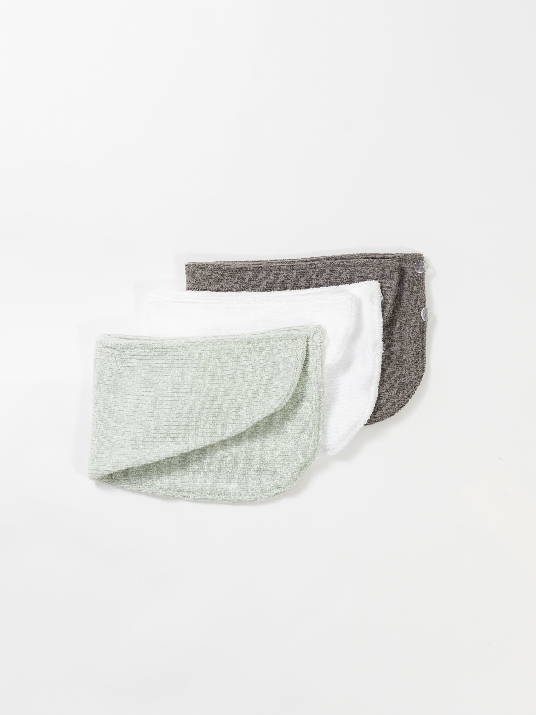 Three folded Ribbed Terry Hair Towels by Cozy Earth are laid out overlapping each other on a white surface. The towels are in soft pastel colors: mint green, white, and charcoal gray. Each towel features a rounded shape with a front flap, and they have snap buttons for fastening. |Color:Charcoal