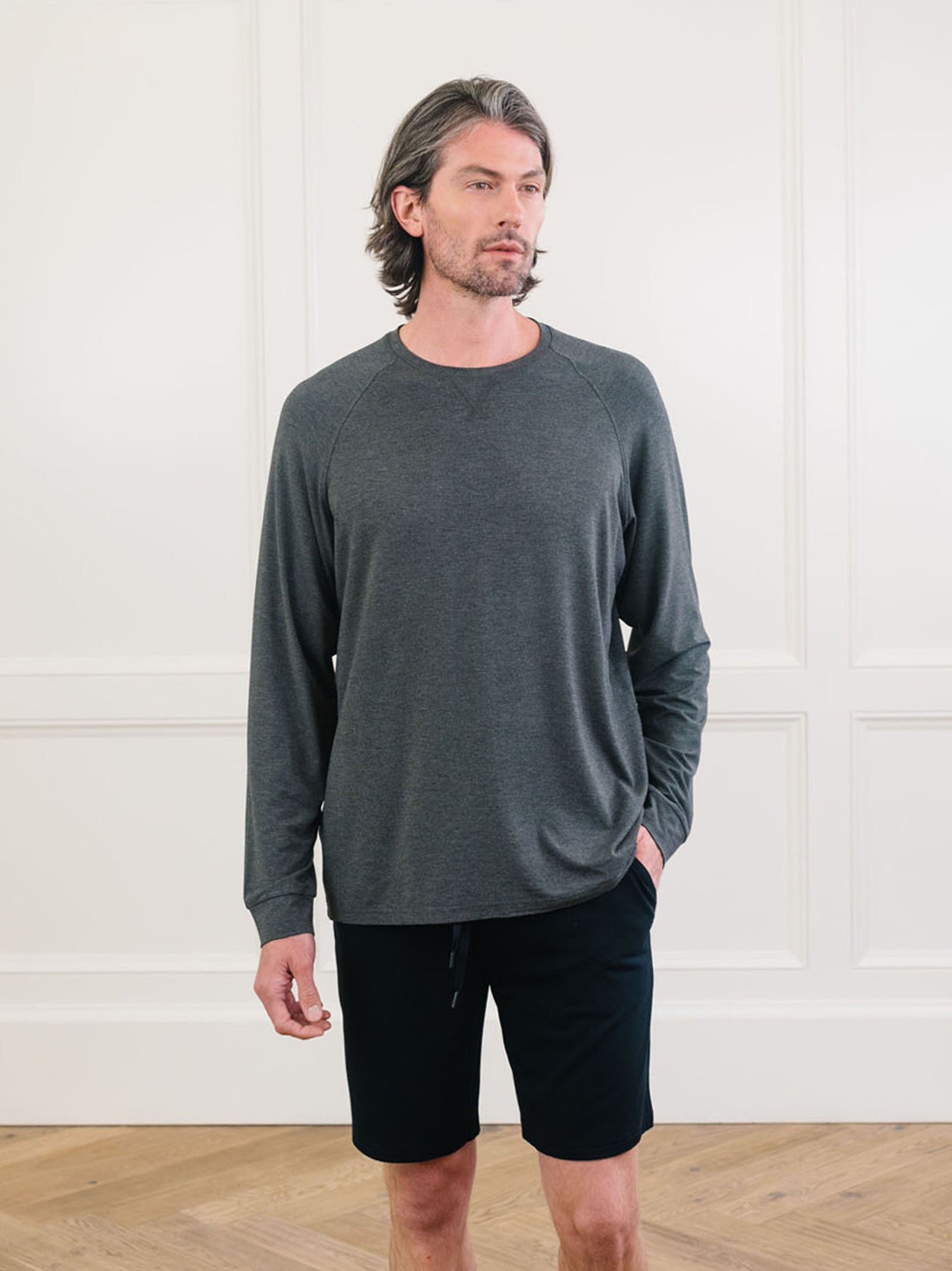 A man with shoulder-length hair is standing in front of a white paneled wall. He is wearing a Cozy Earth Men's Stretch-Knit Long Sleeve Bamboo Pajama Set in dark gray and black shorts, and has his hands in his pockets. The floor beneath him is wooden.  