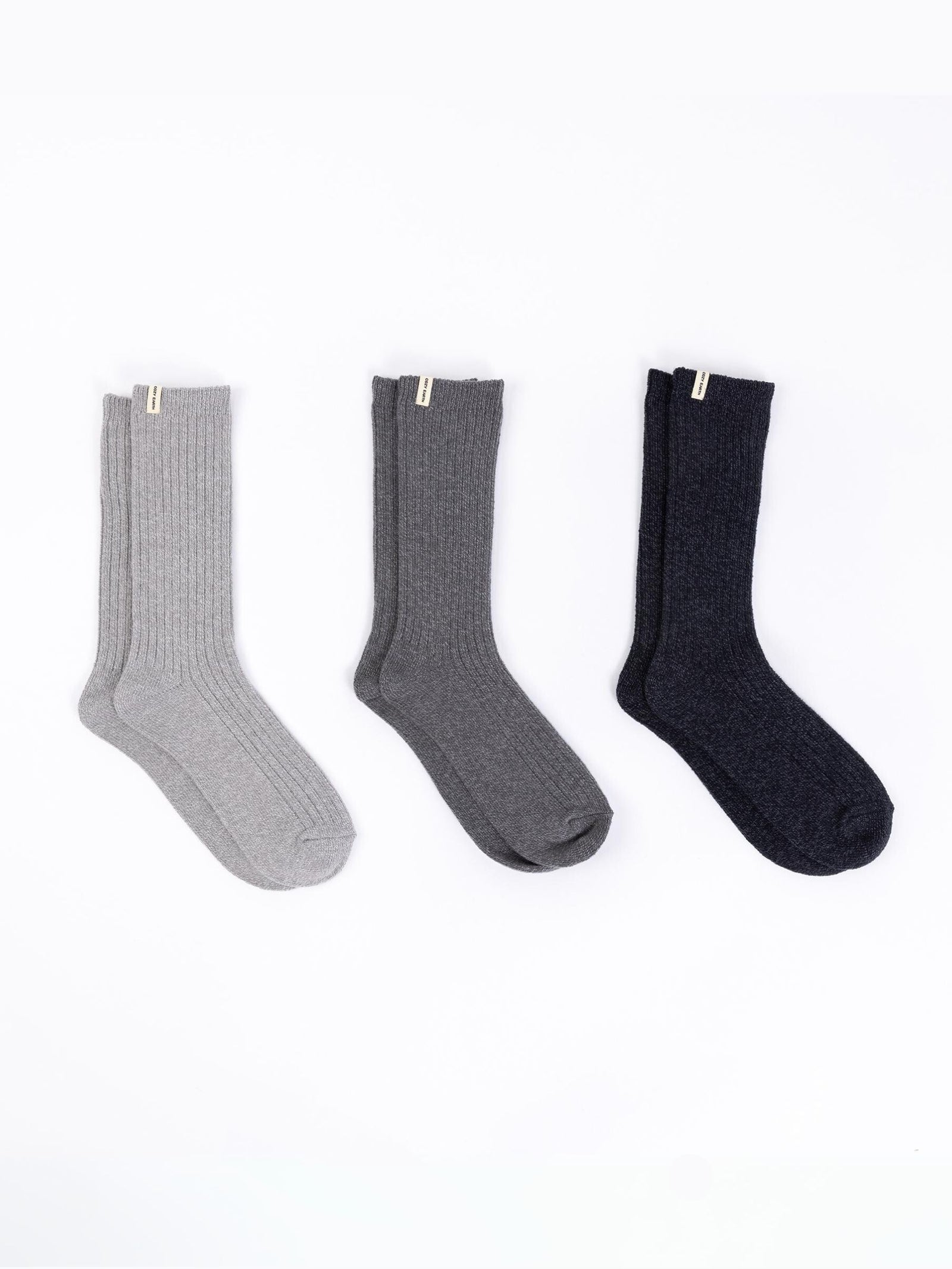 The Plush Lounge Sock 3-Pack from Cozy Earth features three pairs of ribbed socks displayed against a white backdrop, showcasing a palette of light gray, dark gray, and black. Each pair is adorned with a small tag at the top. 