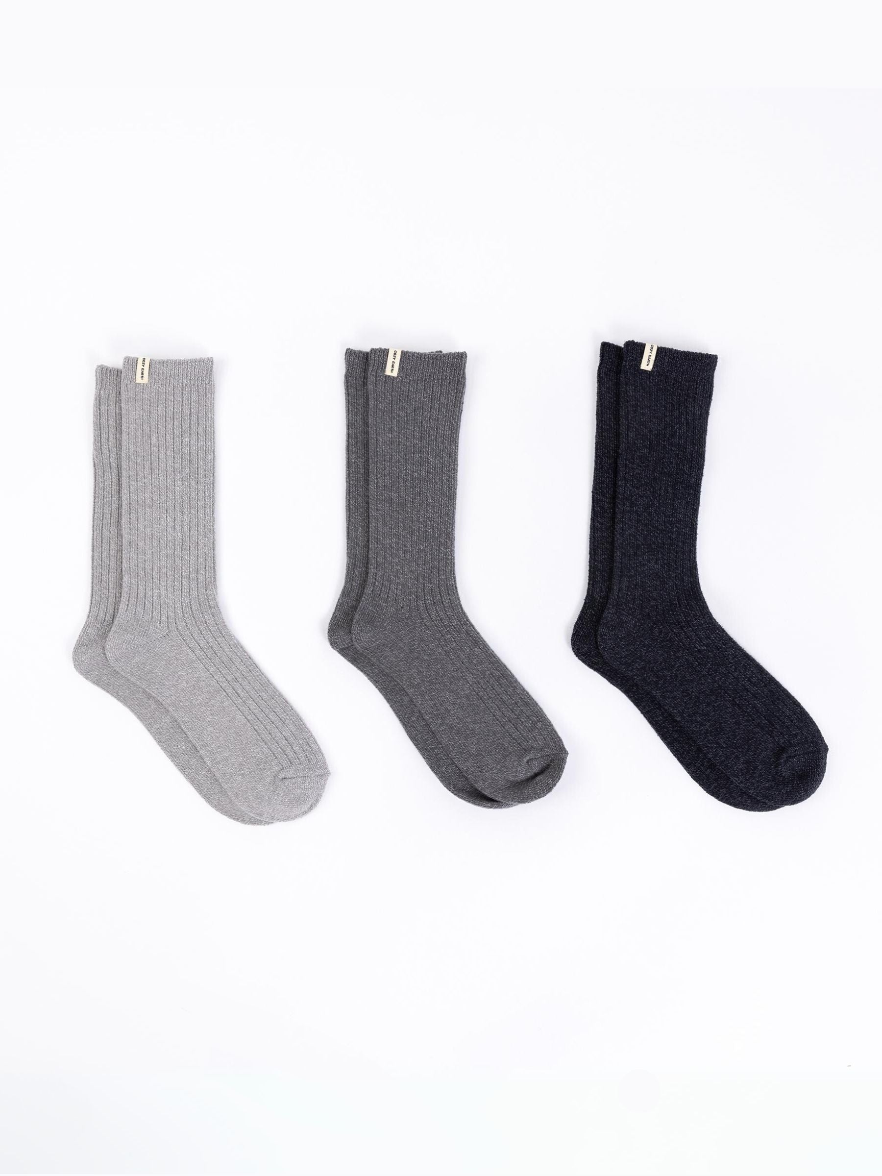 The Plush Lounge Calf Sock 3-Pack from Cozy Earth features three pairs of ribbed socks displayed against a white backdrop, showcasing a palette of light gray, dark gray, and black. Each pair is adorned with a small tag at the top. 