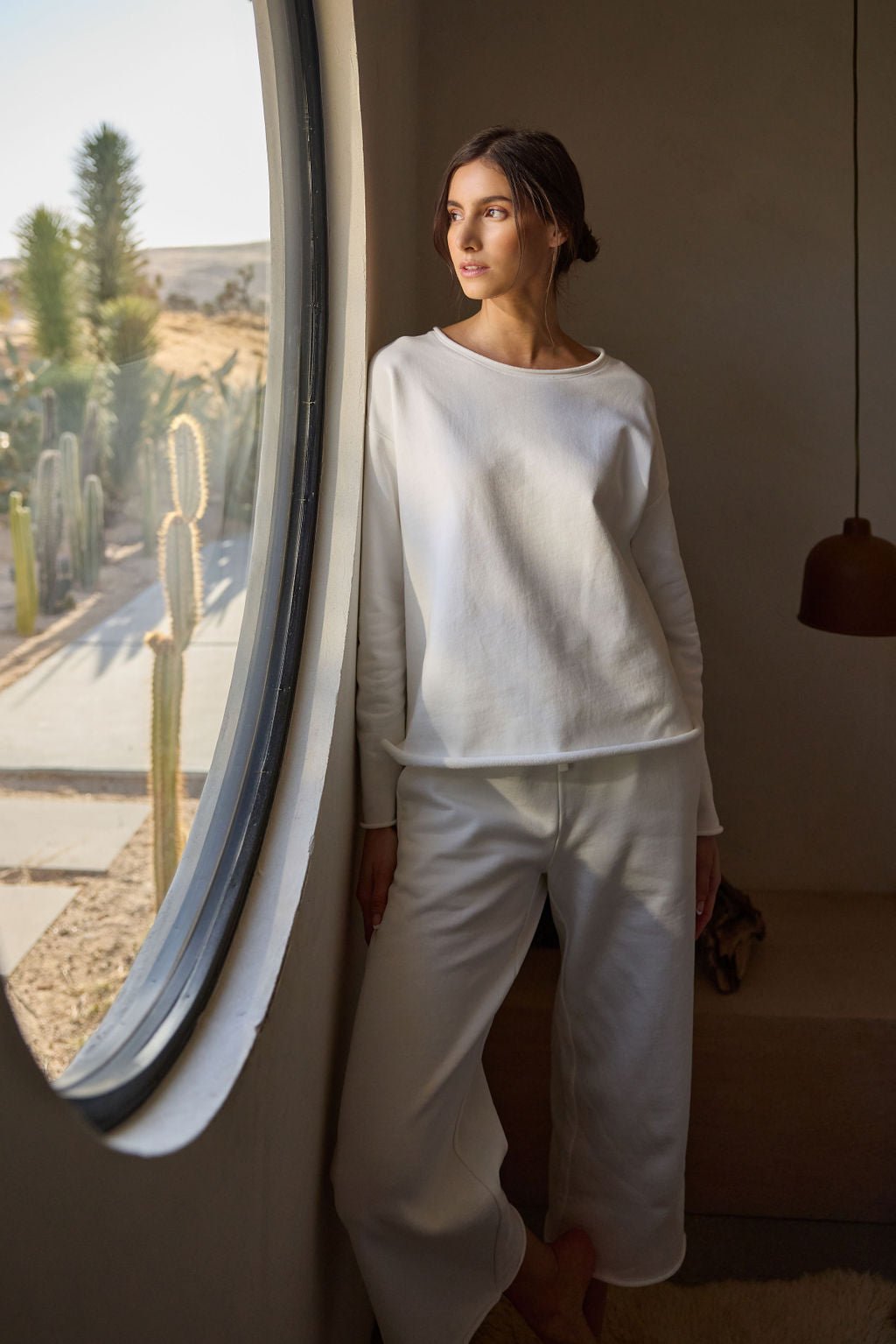 A person wearing Cozy Earth's Women's Chelsea Cropped Pant in white stands by a large round window, gazing at the cacti and desert landscape outside. The softly lit interior features a brown pendant light hanging nearby. 