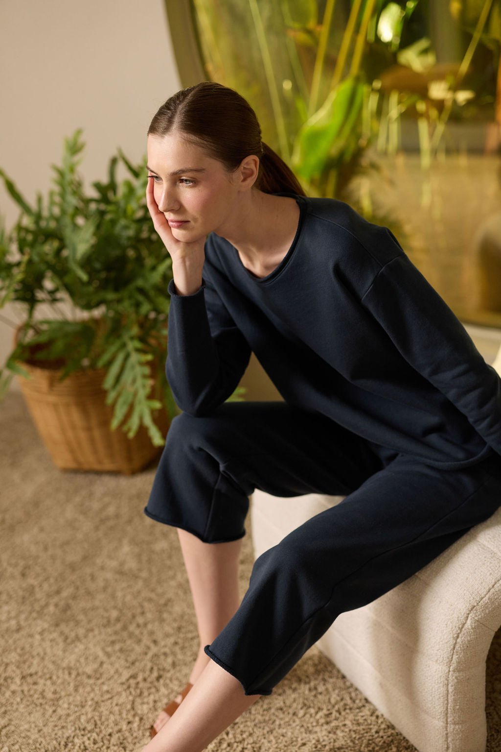 A person with long hair tied back sits on a light bench, wearing a dark, long-sleeved outfit paired with Cozy Earth's Women's Chelsea Cropped Pant. They rest their chin in one hand, gazing sideways. Behind them is a large plant and a circular window. 