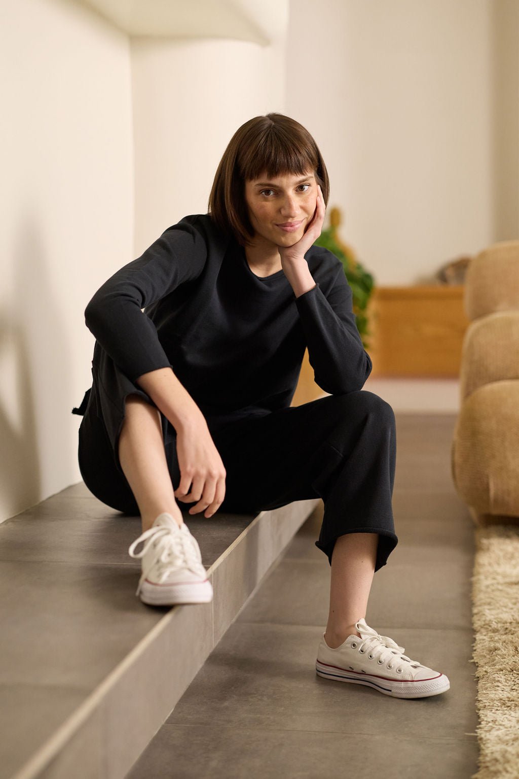 A person with a bob haircut sits on a raised ledge, leaning forward with their chin on one hand, wearing a black outfit and white sneakers. The background shows a beige couch and a plush rug. They are wearing the Women's Chelsea Cropped Pant by Cozy Earth. |Color:Jet Black