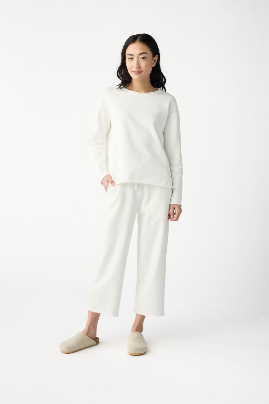 A person models the Women's Chelsea Set by Cozy Earth, featuring a white long-sleeve shirt and matching pants against a plain white background. They have their left hand in their pocket and are wearing beige slippers. 