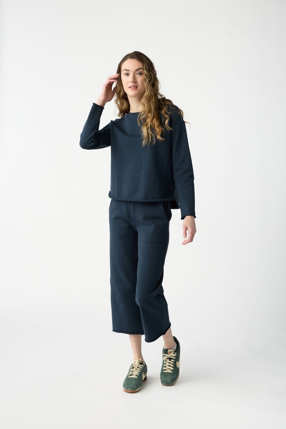 Someone with long hair wears the Women's Chelsea Set by Cozy Earth, a matching dark blue sweatshirt and pants ensemble, paired with green sneakers. They stand against a plain white background, one hand touching their hair. 