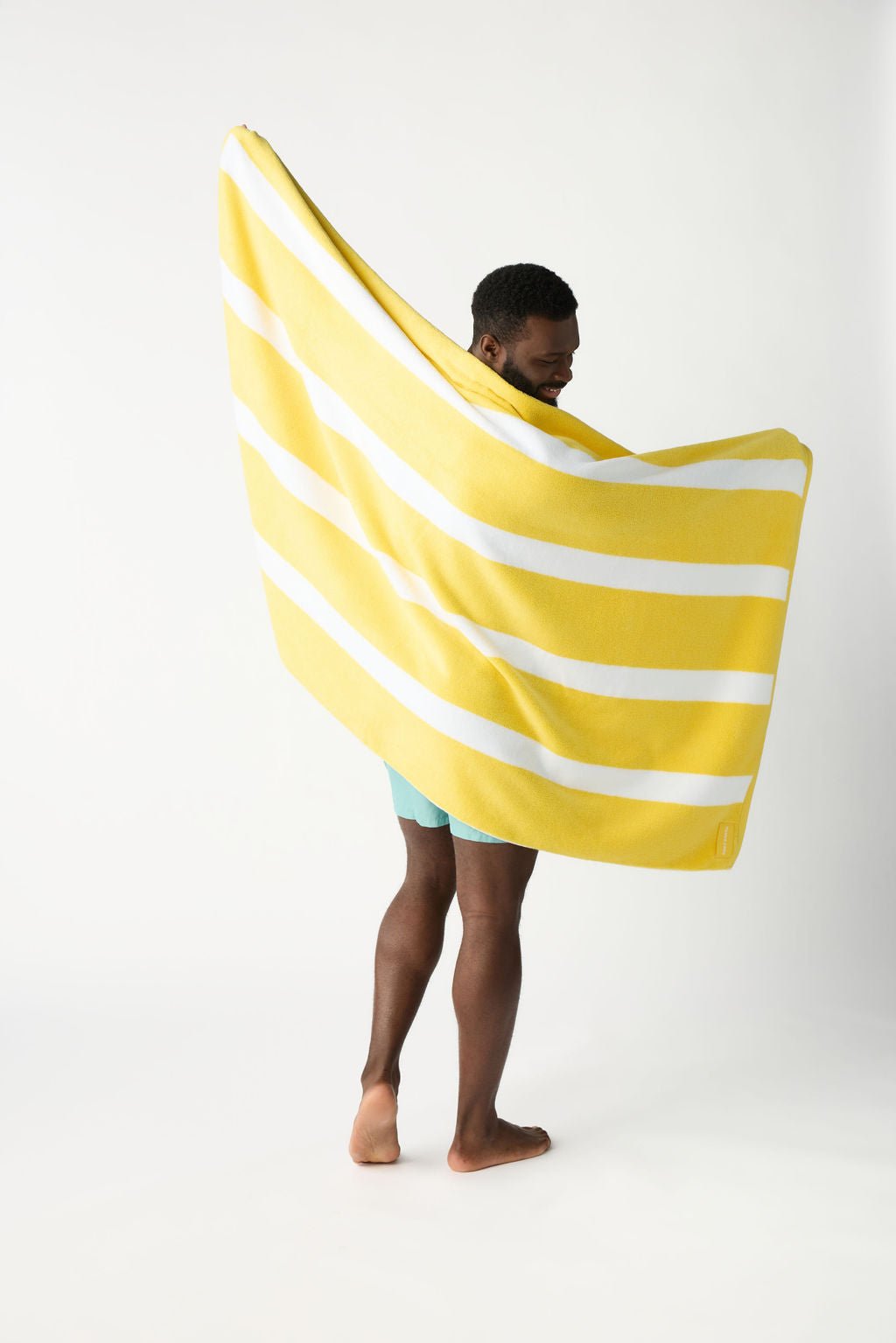 A person balances barefoot on one leg, draping a Classic Stripe Resort Towel from Cozy Earth over their shoulder. They're in blue swim shorts against a plain white background. 
